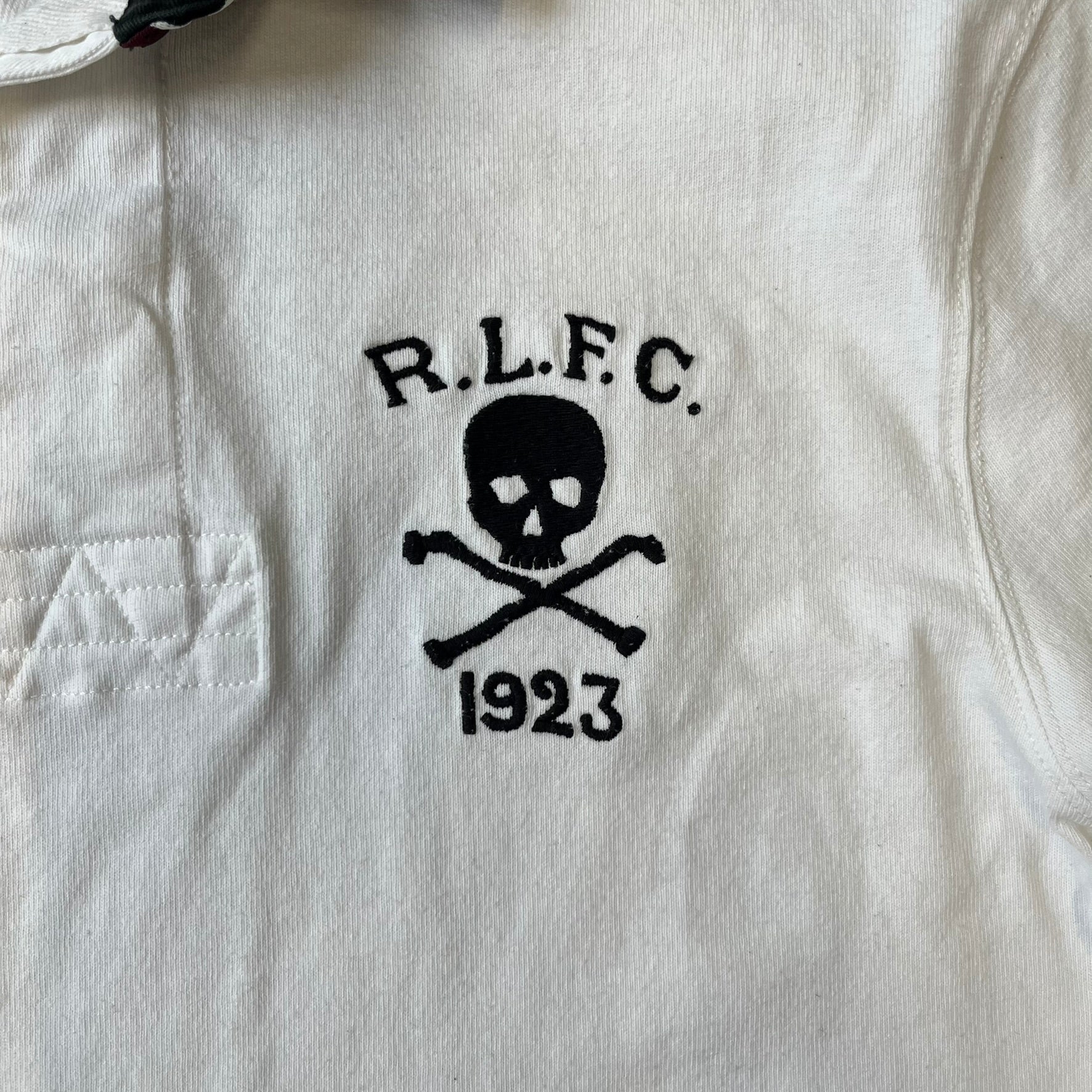 Polo Ralph Lauren “Skull &amp; Crossbone” Rugby Polo is a white short-sleeve polo made from 100% cotton. It features a black contrasting rugby-style collar and a three-button placket. On the chest is a black embroidered skull and crossbones design with “1923” below it and “R.L.F.C” above. The design includes a split hem and embroidered detailing on the front.