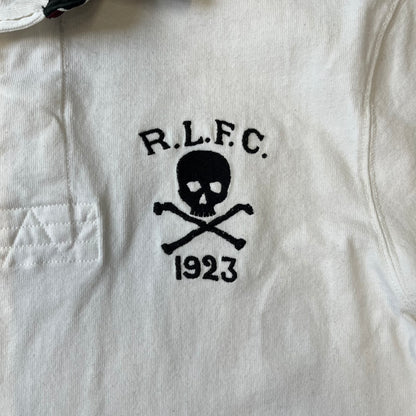 Polo Ralph Lauren “Skull &amp; Crossbone” Rugby Polo is a white short-sleeve polo made from 100% cotton. It features a black contrasting rugby-style collar and a three-button placket. On the chest is a black embroidered skull and crossbones design with “1923” below it and “R.L.F.C” above. The design includes a split hem and embroidered detailing on the front.