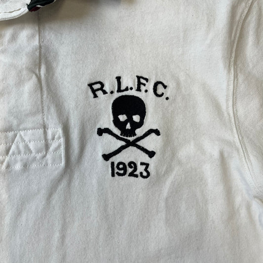 Polo Ralph Lauren “Skull &amp; Crossbone” Rugby Polo is a white short-sleeve polo made from 100% cotton. It features a black contrasting rugby-style collar and a three-button placket. On the chest is a black embroidered skull and crossbones design with “1923” below it and “R.L.F.C” above. The design includes a split hem and embroidered detailing on the front.