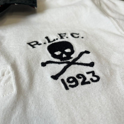 Polo Ralph Lauren “Skull &amp; Crossbone” Rugby Polo is a white short-sleeve polo made from 100% cotton. It features a black contrasting rugby-style collar and a three-button placket. On the chest is a black embroidered skull and crossbones design with “1923” below it and “R.L.F.C” above. The design includes a split hem and embroidered detailing on the front.