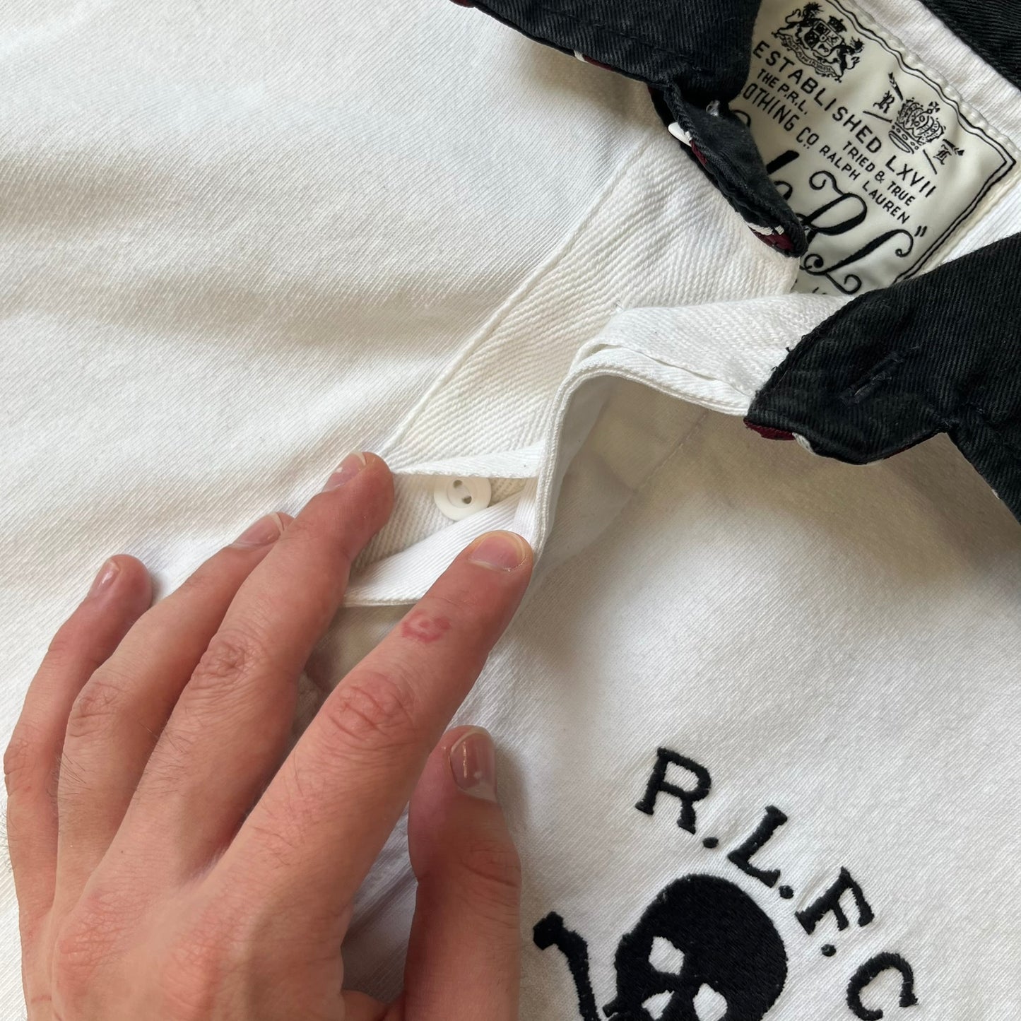 Polo Ralph Lauren “Skull &amp; Crossbone” Rugby Polo is a white short-sleeve polo made from 100% cotton. It features a black contrasting rugby-style collar and a three-button placket. On the chest is a black embroidered skull and crossbones design with “1923” below it and “R.L.F.C” above. The design includes a split hem and embroidered detailing on the front.