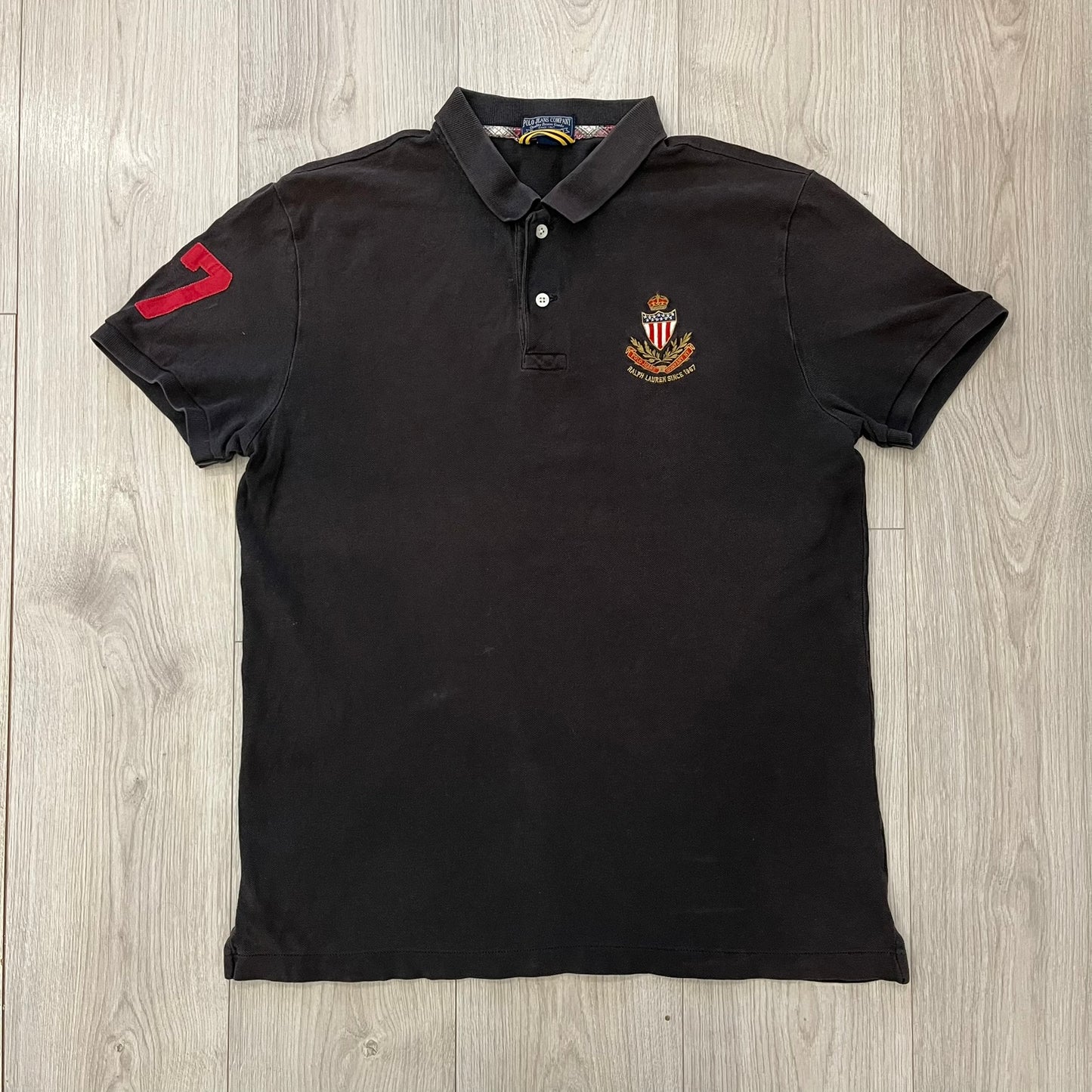 Polo Ralph Lauren ’67 Crest Polo in dark brown. Features an embroidered crest on the left chest with “1967” detail and a big red “67” patch on the sleeve. Classic cotton polo construction with short sleeves and ribbed collar.