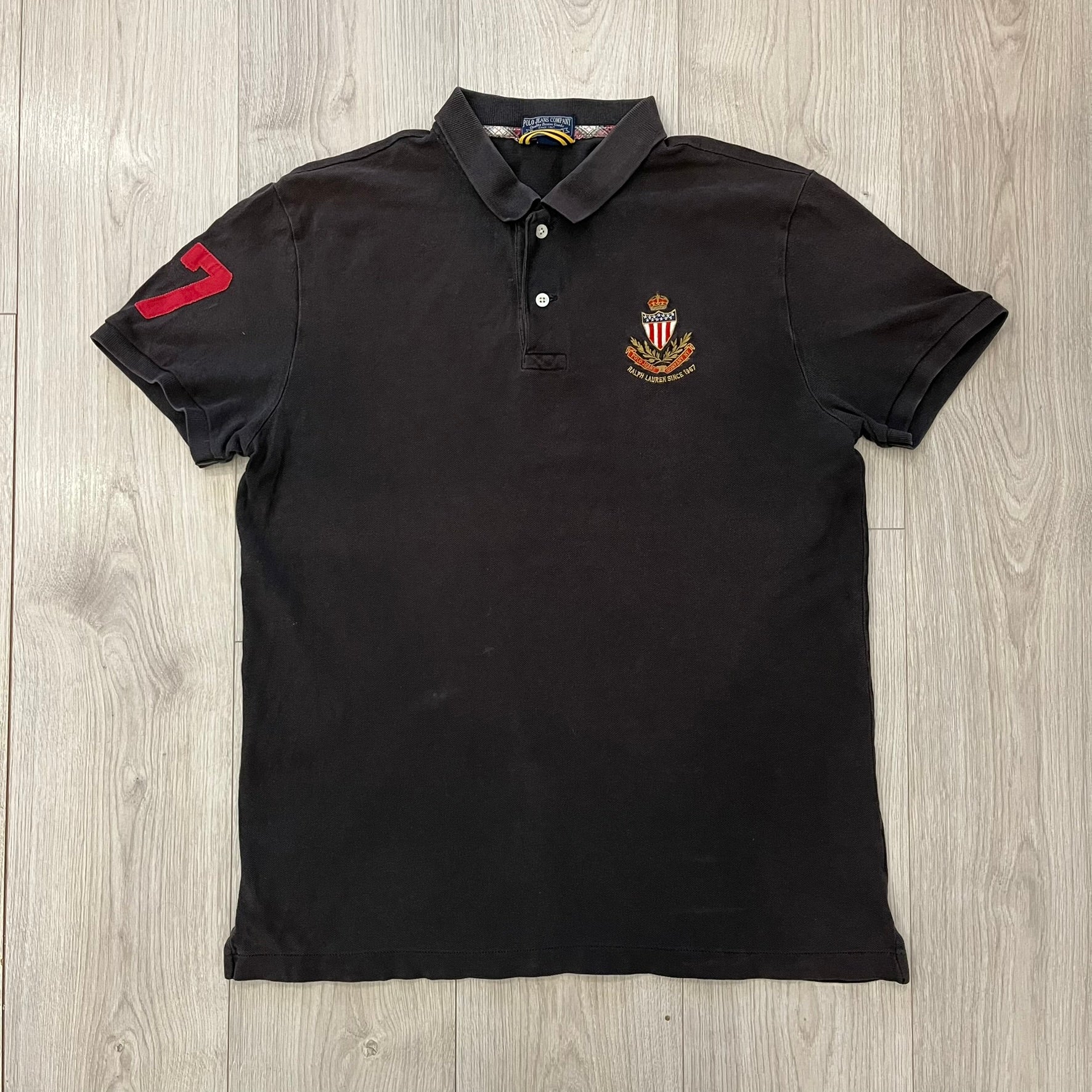 Polo Ralph Lauren ’67 Crest Polo in dark brown. Features an embroidered crest on the left chest with “1967” detail and a big red “67” patch on the sleeve. Classic cotton polo construction with short sleeves and ribbed collar.