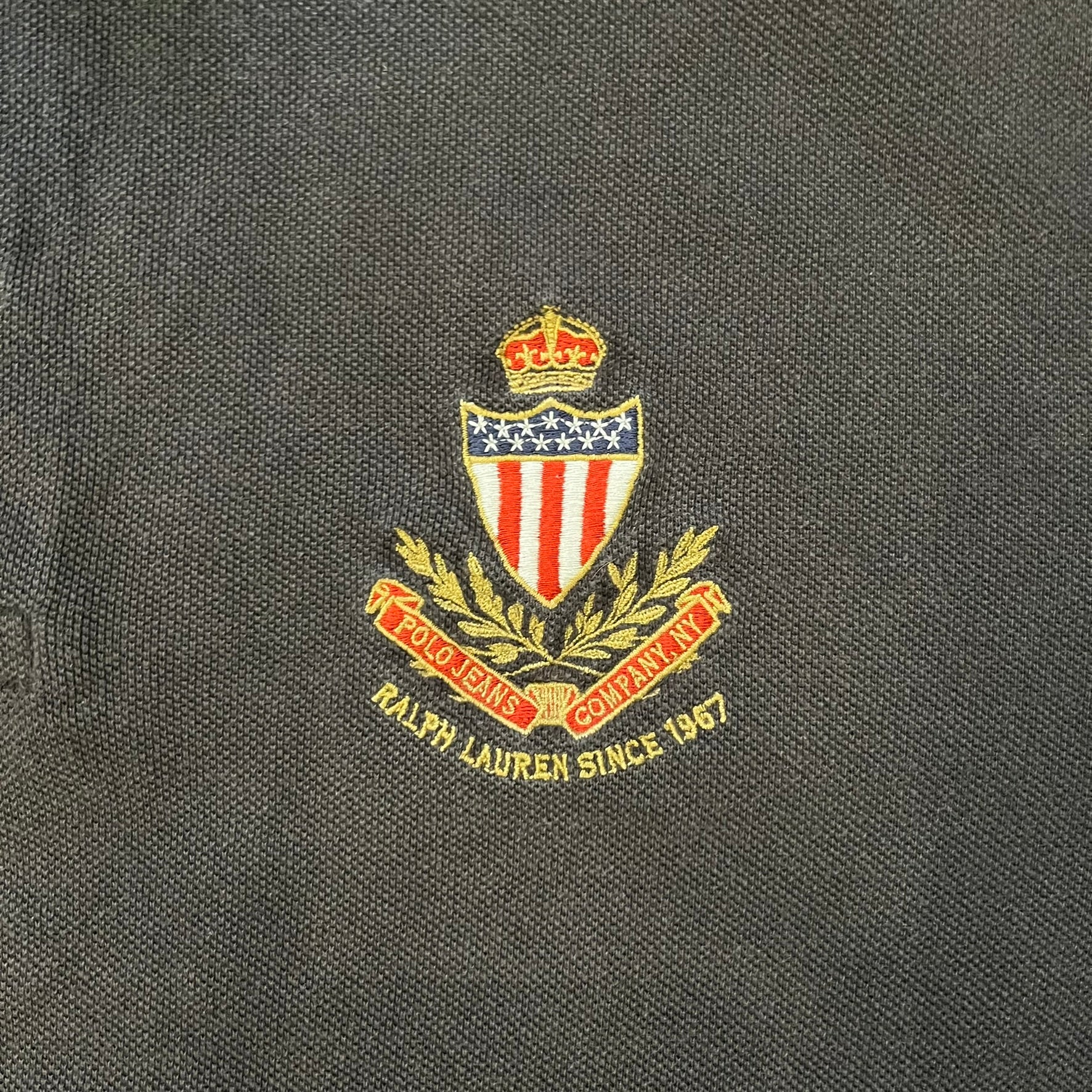 Polo Ralph Lauren ’67 Crest Polo in dark brown. Features an embroidered crest on the left chest with “1967” detail and a big red “67” patch on the sleeve. Classic cotton polo construction with short sleeves and ribbed collar.