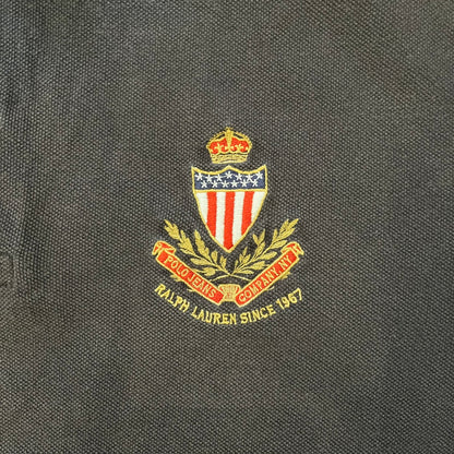 Polo Ralph Lauren ’67 Crest Polo in dark brown. Features an embroidered crest on the left chest with “1967” detail and a big red “67” patch on the sleeve. Classic cotton polo construction with short sleeves and ribbed collar.
