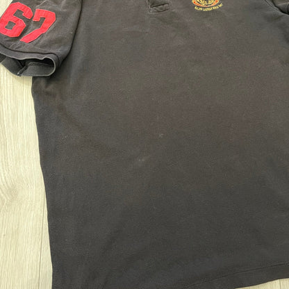Polo Ralph Lauren ’67 Crest Polo in dark brown. Features an embroidered crest on the left chest with “1967” detail and a big red “67” patch on the sleeve. Classic cotton polo construction with short sleeves and ribbed collar.