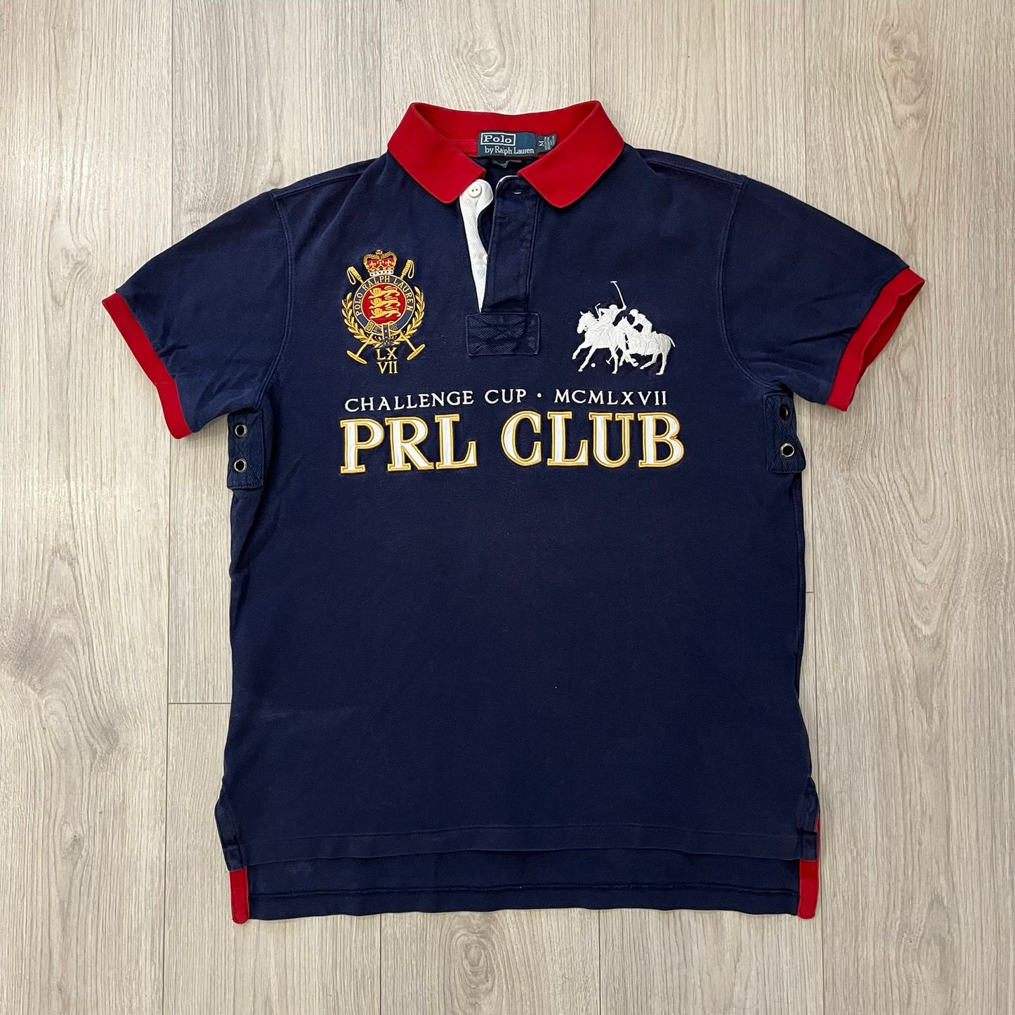 Ralph Lauren’s “PRL Club” Big Pony Polo stands out with detailed front embroidery, combining a Big Pony motif, Challenge Cup text, and a large crest. The navy body is paired with red accents on the collar and sleeves, with a signature oversized “3” and PRL Club branding on the back.