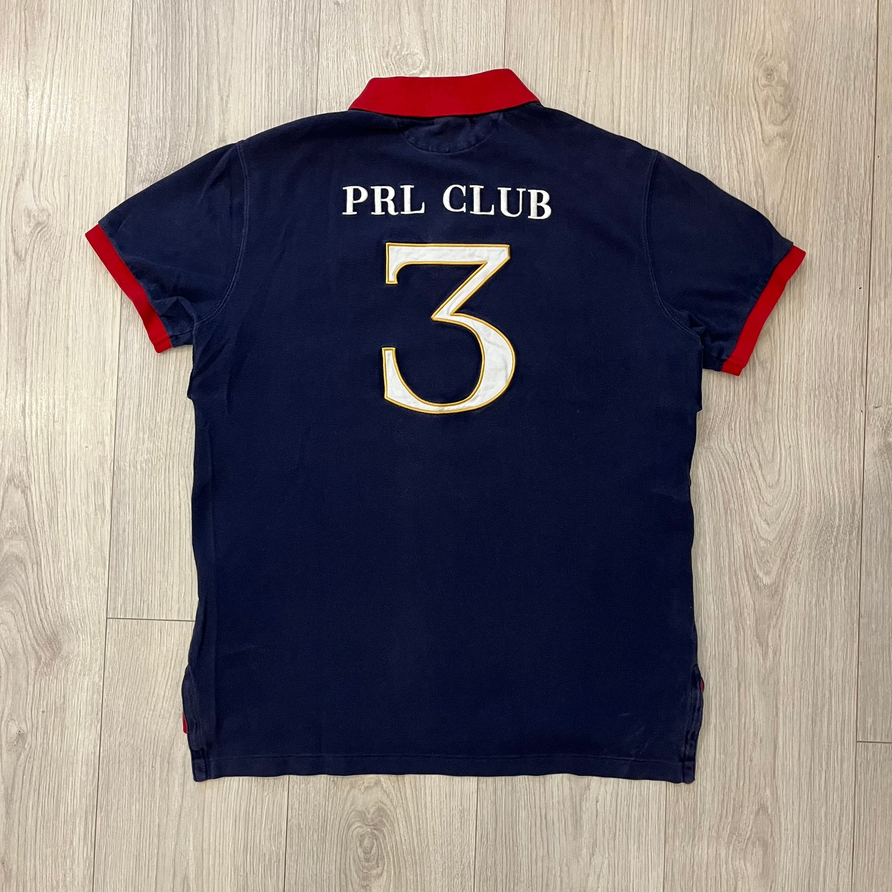Ralph Lauren’s “PRL Club” Big Pony Polo stands out with detailed front embroidery, combining a Big Pony motif, Challenge Cup text, and a large crest. The navy body is paired with red accents on the collar and sleeves, with a signature oversized “3” and PRL Club branding on the back.