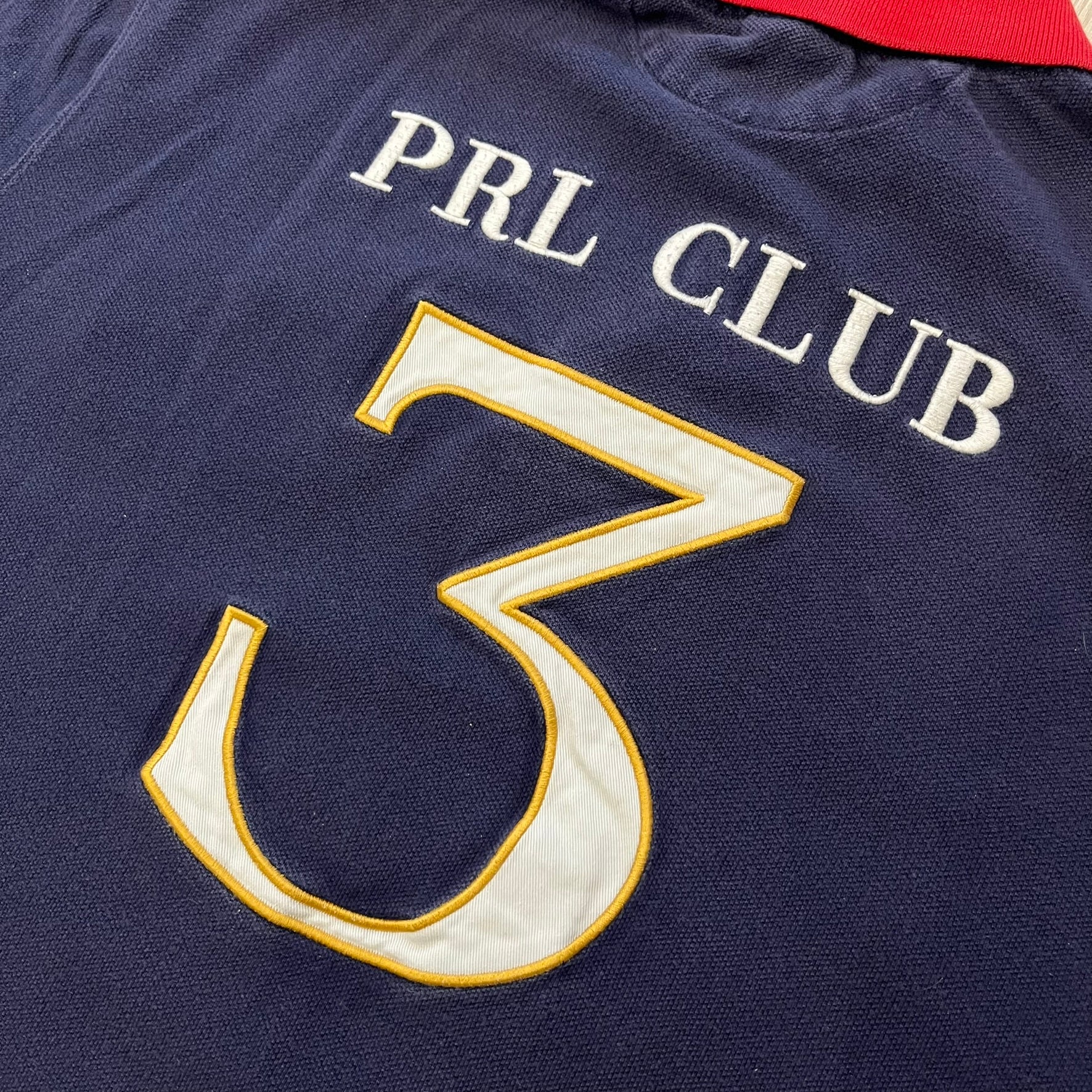 Ralph Lauren’s “PRL Club” Big Pony Polo stands out with detailed front embroidery, combining a Big Pony motif, Challenge Cup text, and a large crest. The navy body is paired with red accents on the collar and sleeves, with a signature oversized “3” and PRL Club branding on the back.
