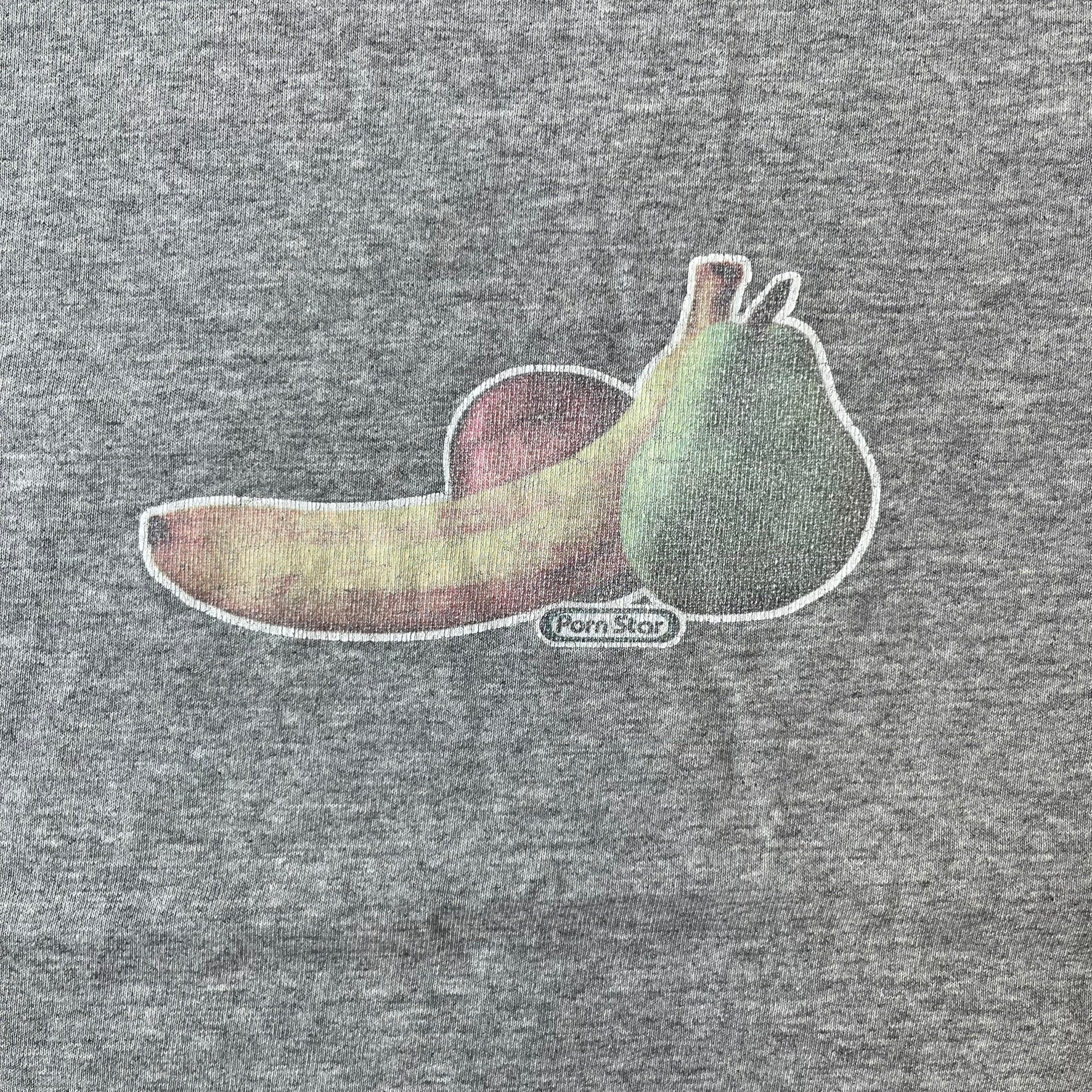 Porn Star Clothing 90's Fruits Gray Tee features a grey T-shirt with an apple, banana, and pear graphic on the chest, arranged as a you know what. A small Porn Star logo is printed underneath, with the “Good Clothing for Bad People” branding on the neck tag. The shirt has two professionally repaired holes under the armpits.