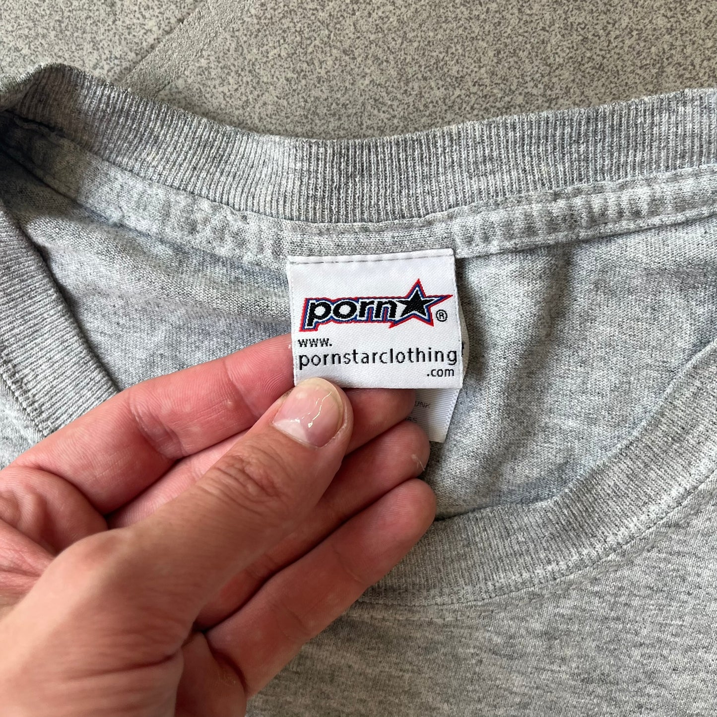 Porn Star Clothing 90's Fruits Gray Tee features a grey T-shirt with an apple, banana, and pear graphic on the chest, arranged as a you know what. A small Porn Star logo is printed underneath, with the “Good Clothing for Bad People” branding on the neck tag. The shirt has two professionally repaired holes under the armpits.