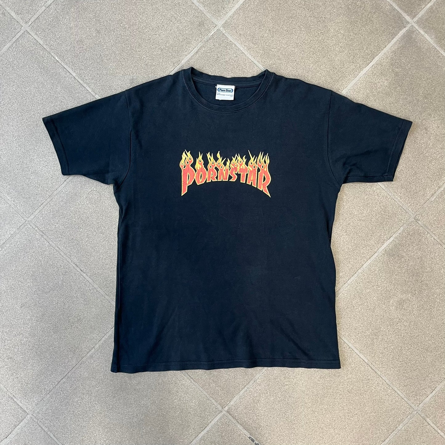 Porn Star Clothing 90s Trasher Black Tee features a black heavyweight cotton construction with a red flaming “Pornstar” logo graphic on the front in a design inspired by Trasher-style branding. The tee has a Porn Star neck tag and is made in the USA.