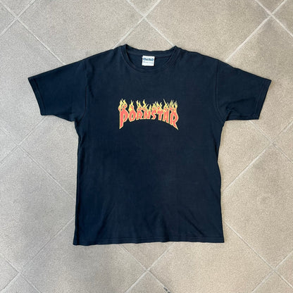 Porn Star Clothing 90s Trasher Black Tee features a black heavyweight cotton construction with a red flaming “Pornstar” logo graphic on the front in a design inspired by Trasher-style branding. The tee has a Porn Star neck tag and is made in the USA.