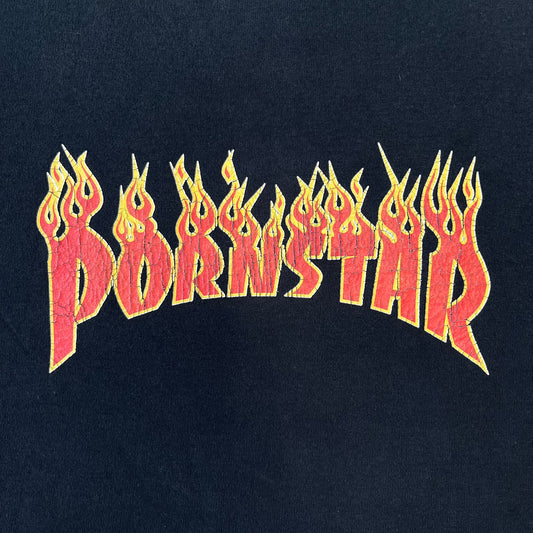 Porn Star Clothing 90s Trasher Black Tee features a black heavyweight cotton construction with a red flaming “Pornstar” logo graphic on the front in a design inspired by Trasher-style branding. The tee has a Porn Star neck tag and is made in the USA.