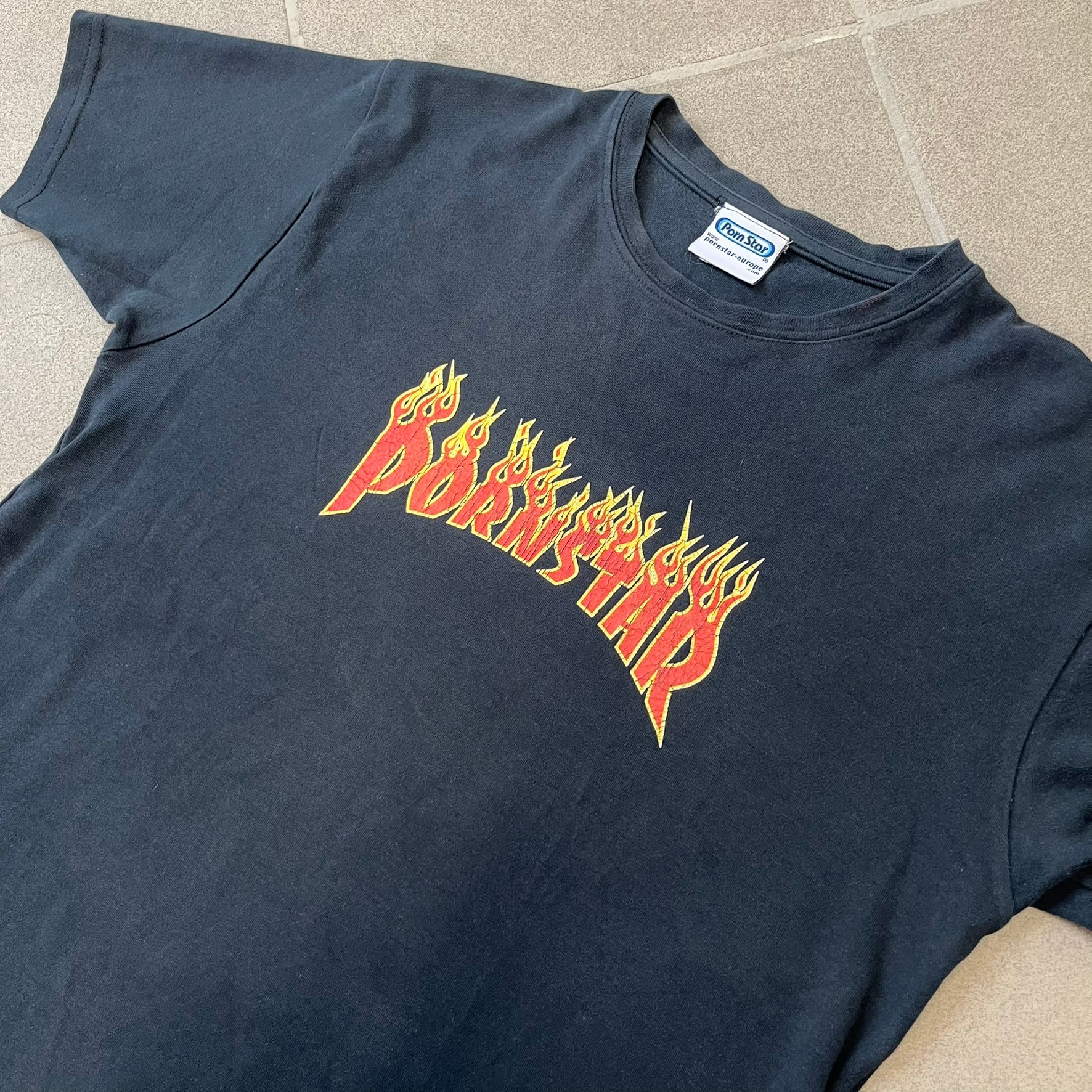 Porn Star Clothing 90s Trasher Black Tee features a black heavyweight cotton construction with a red flaming “Pornstar” logo graphic on the front in a design inspired by Trasher-style branding. The tee has a Porn Star neck tag and is made in the USA.