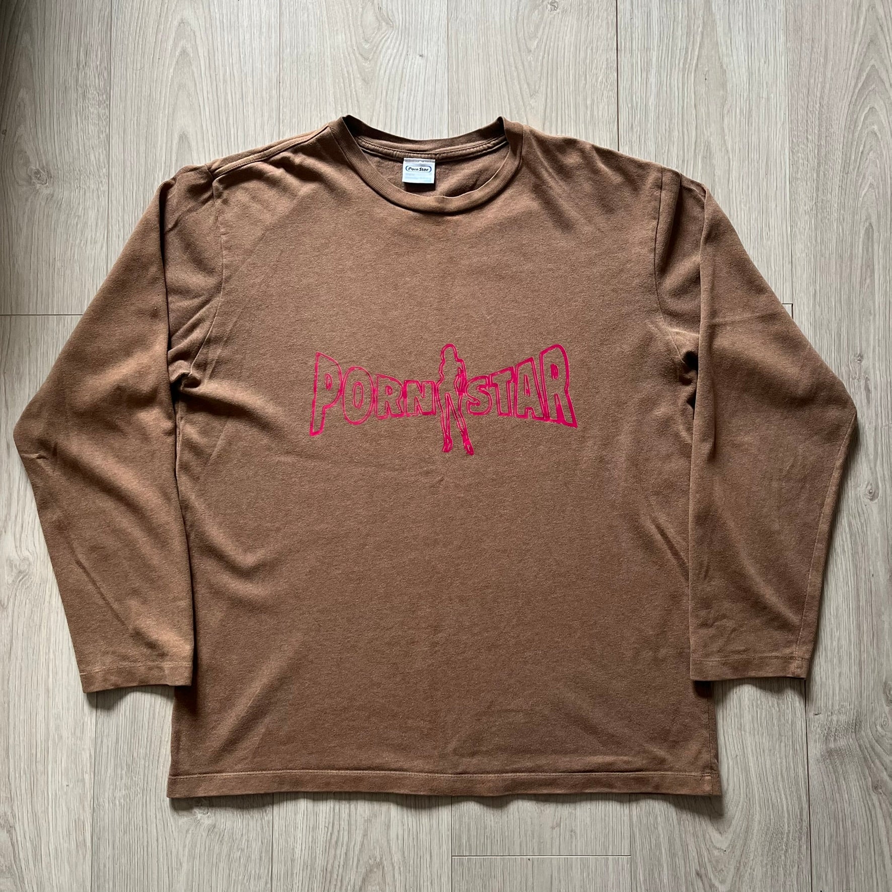 Vintage 90’s Porn Star Clothing longsleeve in brown, featuring a bold pink logo on the chest with a silhouette outline of a woman between the words “Porn” and “Star.” Crafted with quality construction, it carries the brand’s signature edgy aesthetic. Finished with a playful “must be 18 or over” detail on the tag, this rare piece embodies 90’s streetwear provocative style.