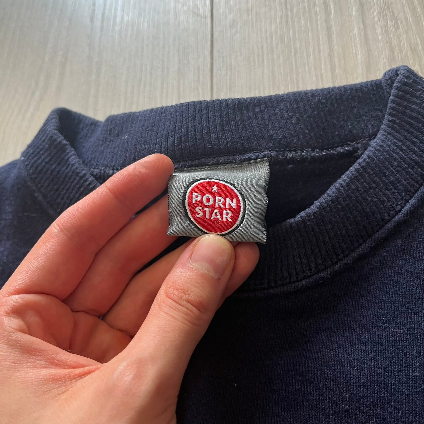 Navy blue sweater with a 'Porn Star' label held by a hand on a wooden surface.