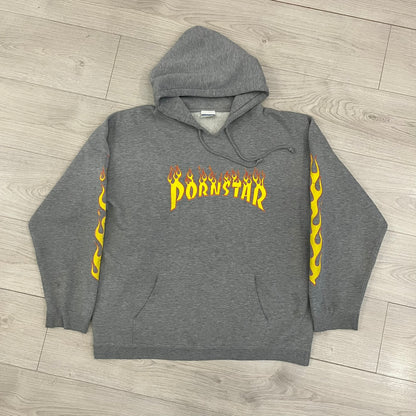 Porn Star Clothing Thrasher Hoodie from the 1990s, made in a gray cotton fabric. The front features a large flame-style “Pornstar” logo that is a spin-off of the Trasher one, with matching flame graphics running down both sleeves. Designed with an adjustable drawstring hood, front kangaroo pocket, and ribbed cuffs and hem. Finished with the original “Must Be 18 Or Over” Porn Star Clothing tag.