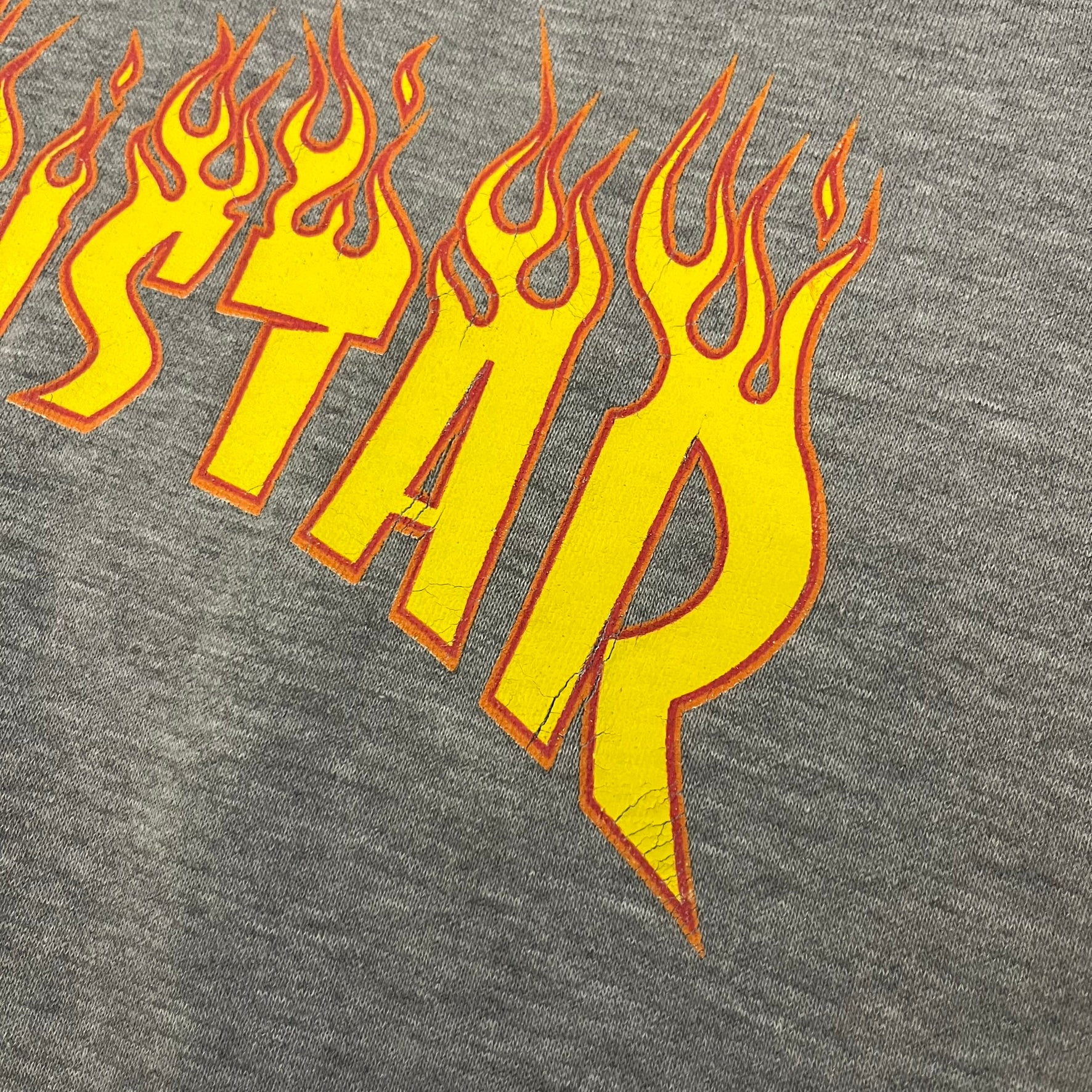 Porn Star Clothing Thrasher Hoodie from the 1990s, made in a gray cotton fabric. The front features a large flame-style “Pornstar” logo that is a spin-off of the Trasher one, with matching flame graphics running down both sleeves. Designed with an adjustable drawstring hood, front kangaroo pocket, and ribbed cuffs and hem. Finished with the original “Must Be 18 Or Over” Porn Star Clothing tag.