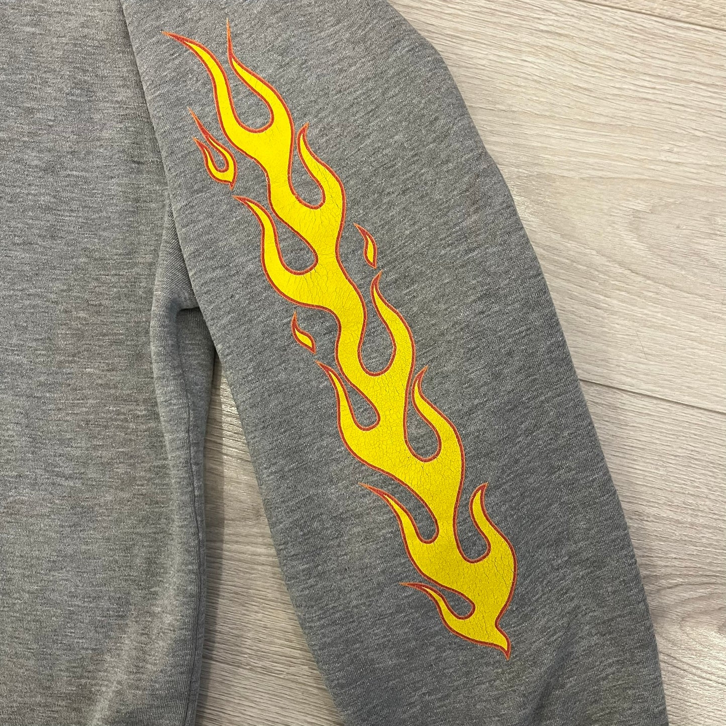 Porn Star Clothing Thrasher Hoodie from the 1990s, made in a gray cotton fabric. The front features a large flame-style “Pornstar” logo that is a spin-off of the Trasher one, with matching flame graphics running down both sleeves. Designed with an adjustable drawstring hood, front kangaroo pocket, and ribbed cuffs and hem. Finished with the original “Must Be 18 Or Over” Porn Star Clothing tag.