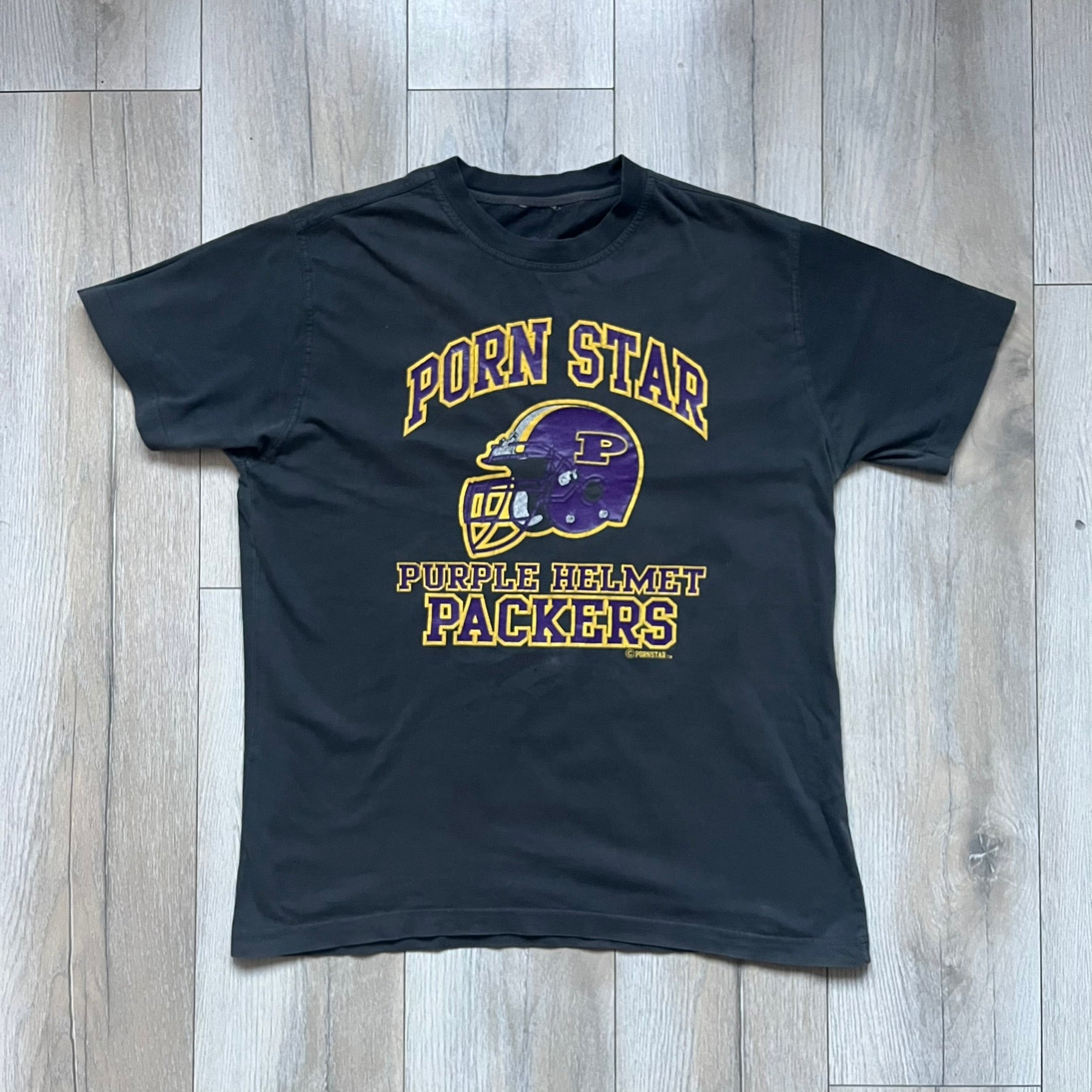Black T-shirt with a purple football helmet graphic featuring a “P” letter. “Porn Star” text above the helmet in purple with a yellow outline, and “Helmet Packers” text below in purple with a yellow outline. Includes a small Porn Star copyright logo. Vintage piece.