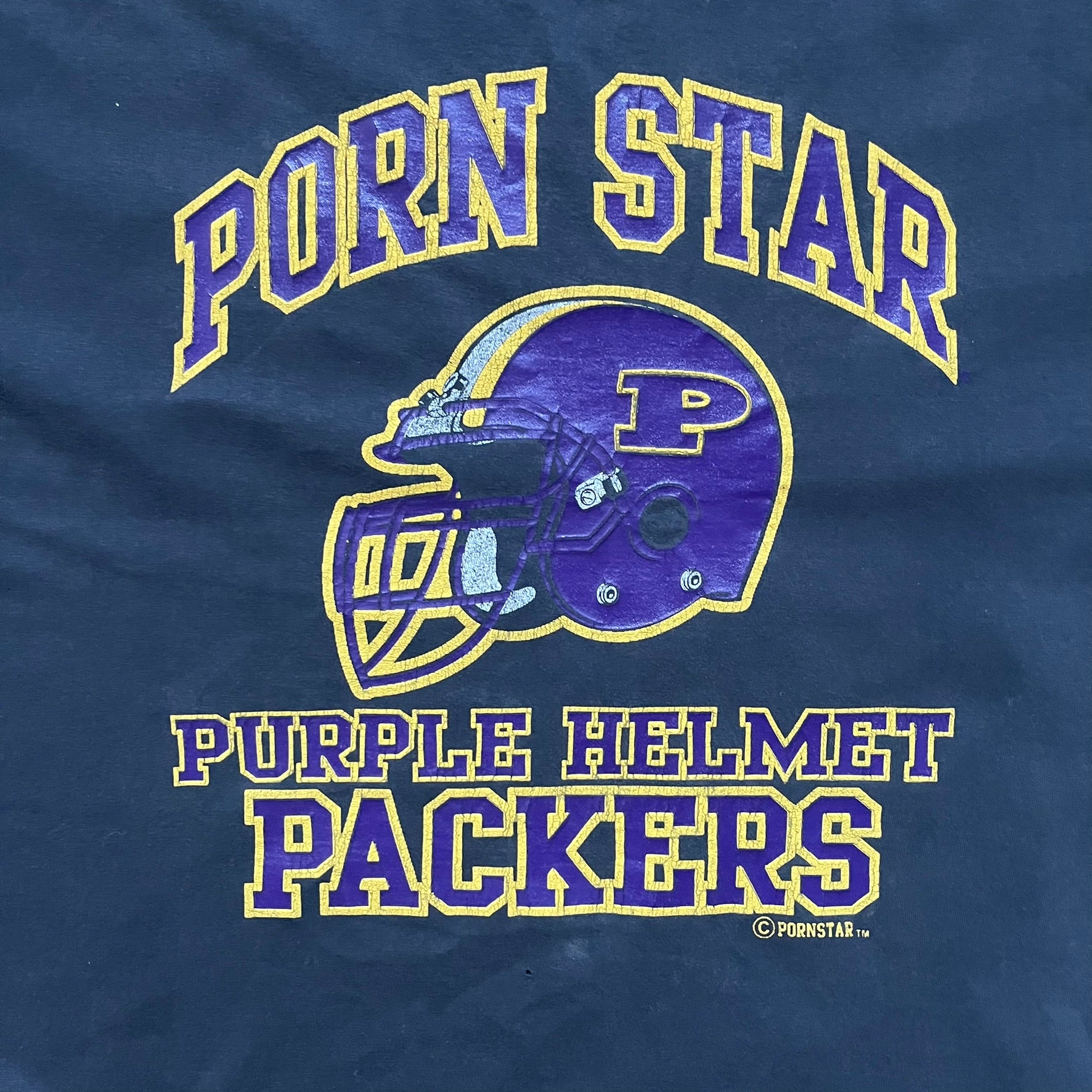 Black T-shirt with a purple football helmet graphic featuring a “P” letter. “Porn Star” text above the helmet in purple with a yellow outline, and “Helmet Packers” text below in purple with a yellow outline. Includes a small Porn Star copyright logo. Vintage piece.