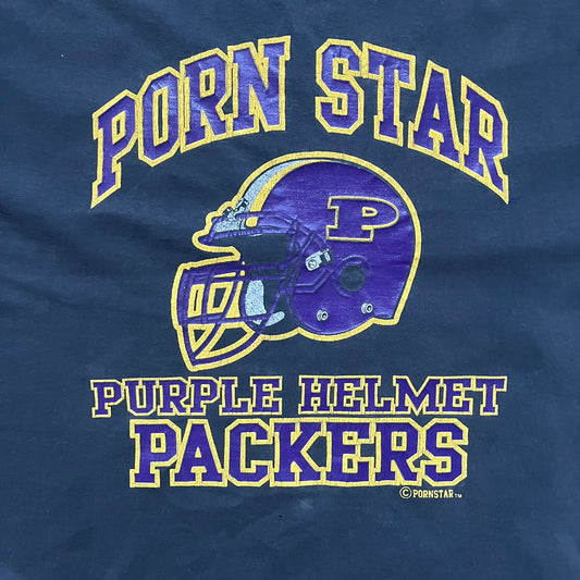 Black T-shirt with a purple football helmet graphic featuring a “P” letter. “Porn Star” text above the helmet in purple with a yellow outline, and “Helmet Packers” text below in purple with a yellow outline. Includes a small Porn Star copyright logo. Vintage piece.