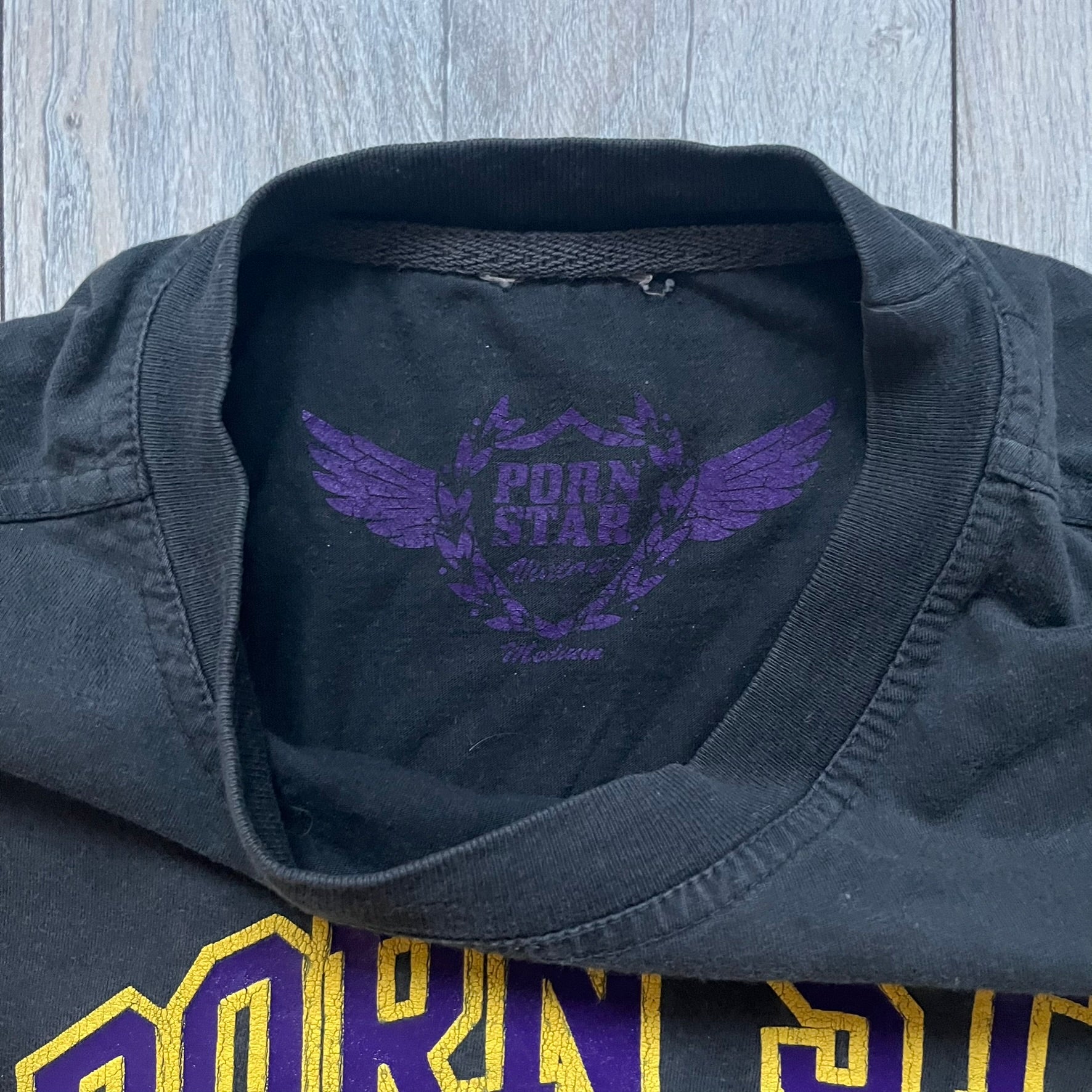 Black T-shirt with a purple football helmet graphic featuring a “P” letter. “Porn Star” text above the helmet in purple with a yellow outline, and “Helmet Packers” text below in purple with a yellow outline. Includes a small Porn Star copyright logo. Vintage piece.