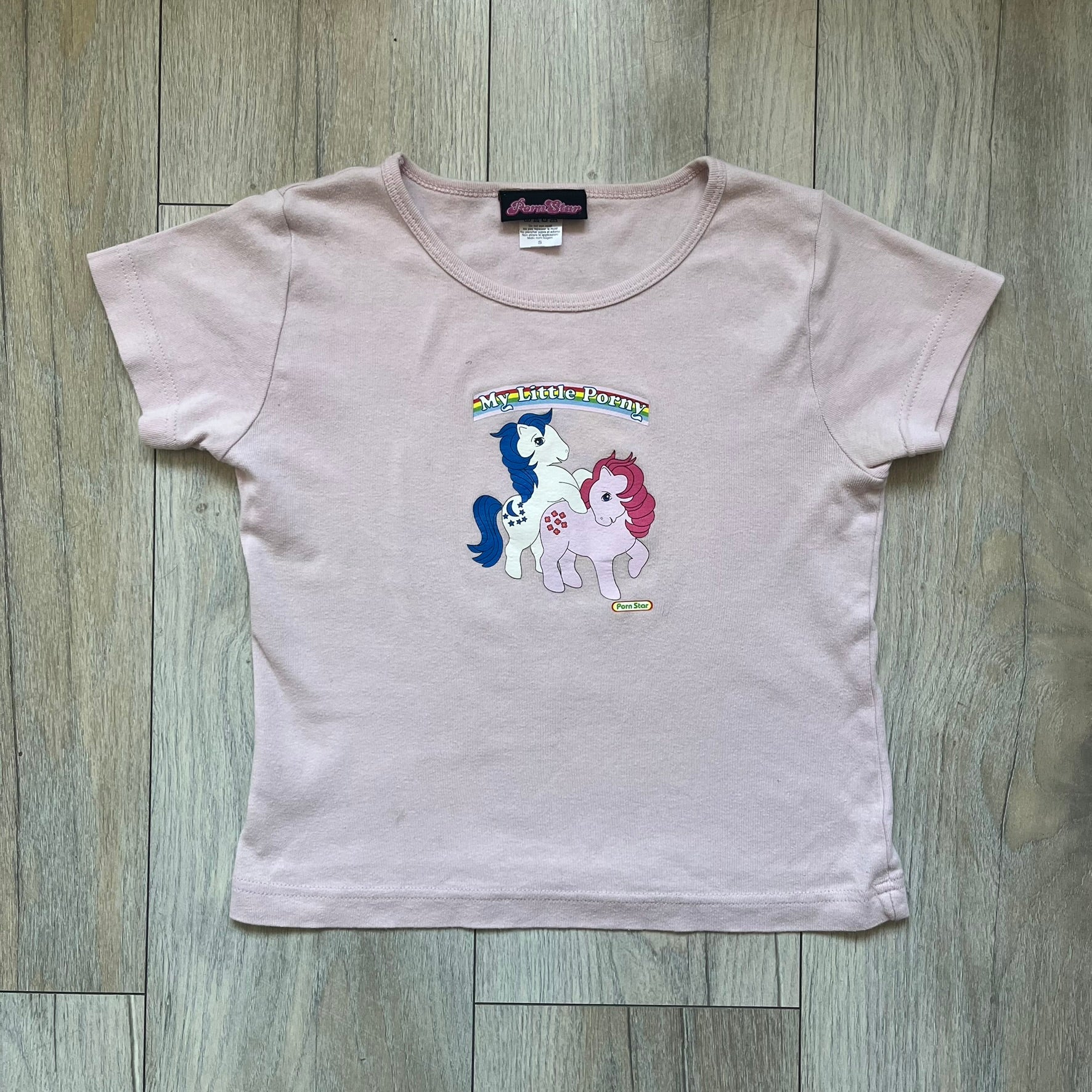 Pornstar Clothing My Little Pony Pink Tee is a women’s baby pink T-shirt featuring a front graphic of two My Little Pony characters under a “My Little Porny” text on a rainbow. A small Pornstar logo sits below the graphic, and the neck tag is black and pink with Pornstar branding. Super rare and hard to find piece.