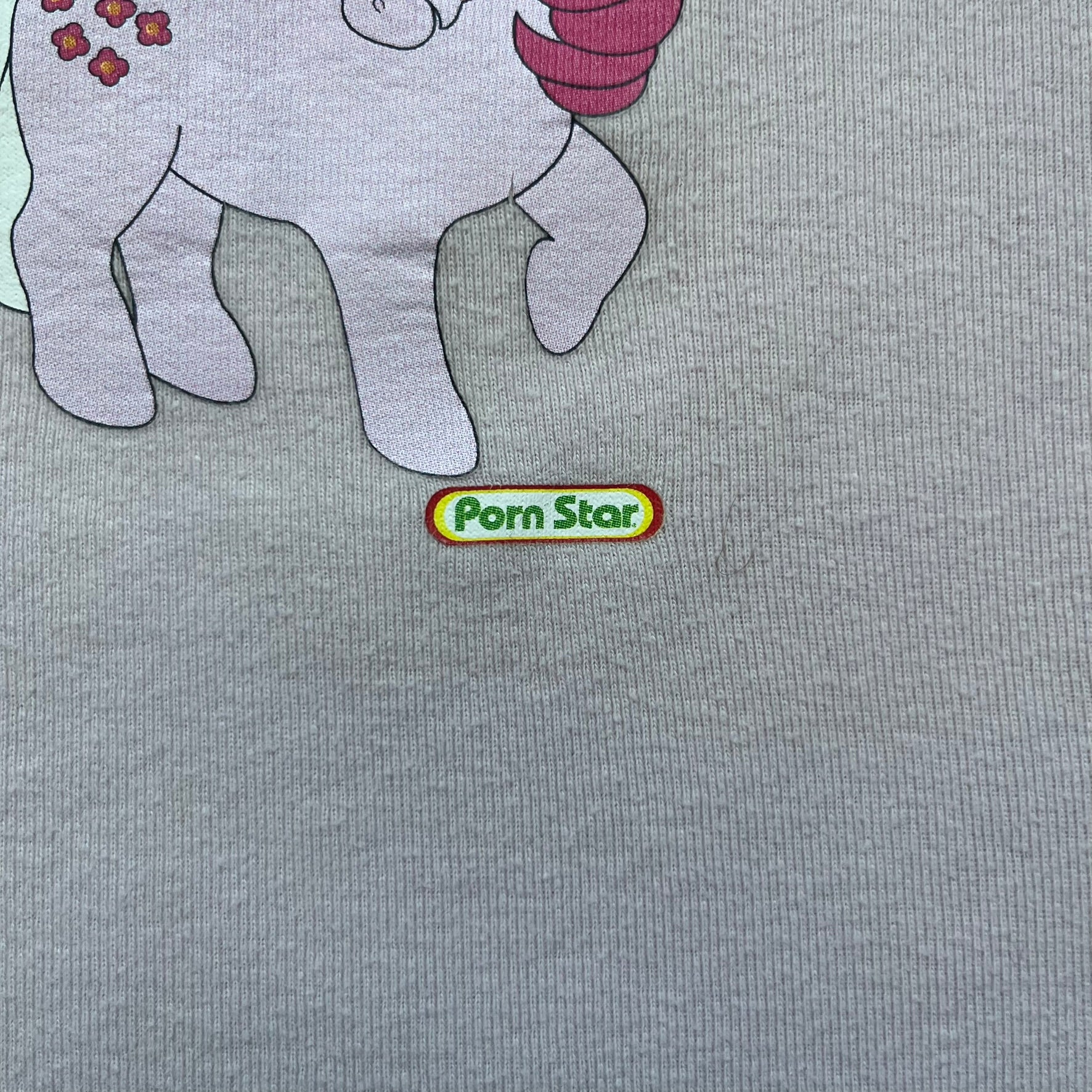 Pornstar Clothing My Little Pony Pink Tee is a women’s baby pink T-shirt featuring a front graphic of two My Little Pony characters under a “My Little Porny” text on a rainbow. A small Pornstar logo sits below the graphic, and the neck tag is black and pink with Pornstar branding. Super rare and hard to find piece.