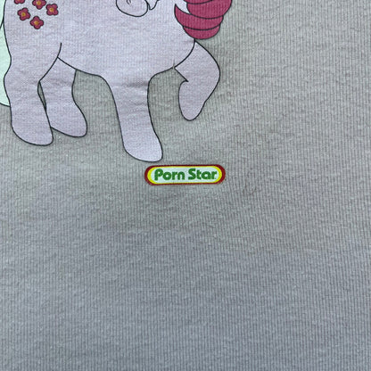 Pornstar Clothing My Little Pony Pink Tee is a women’s baby pink T-shirt featuring a front graphic of two My Little Pony characters under a “My Little Porny” text on a rainbow. A small Pornstar logo sits below the graphic, and the neck tag is black and pink with Pornstar branding. Super rare and hard to find piece.