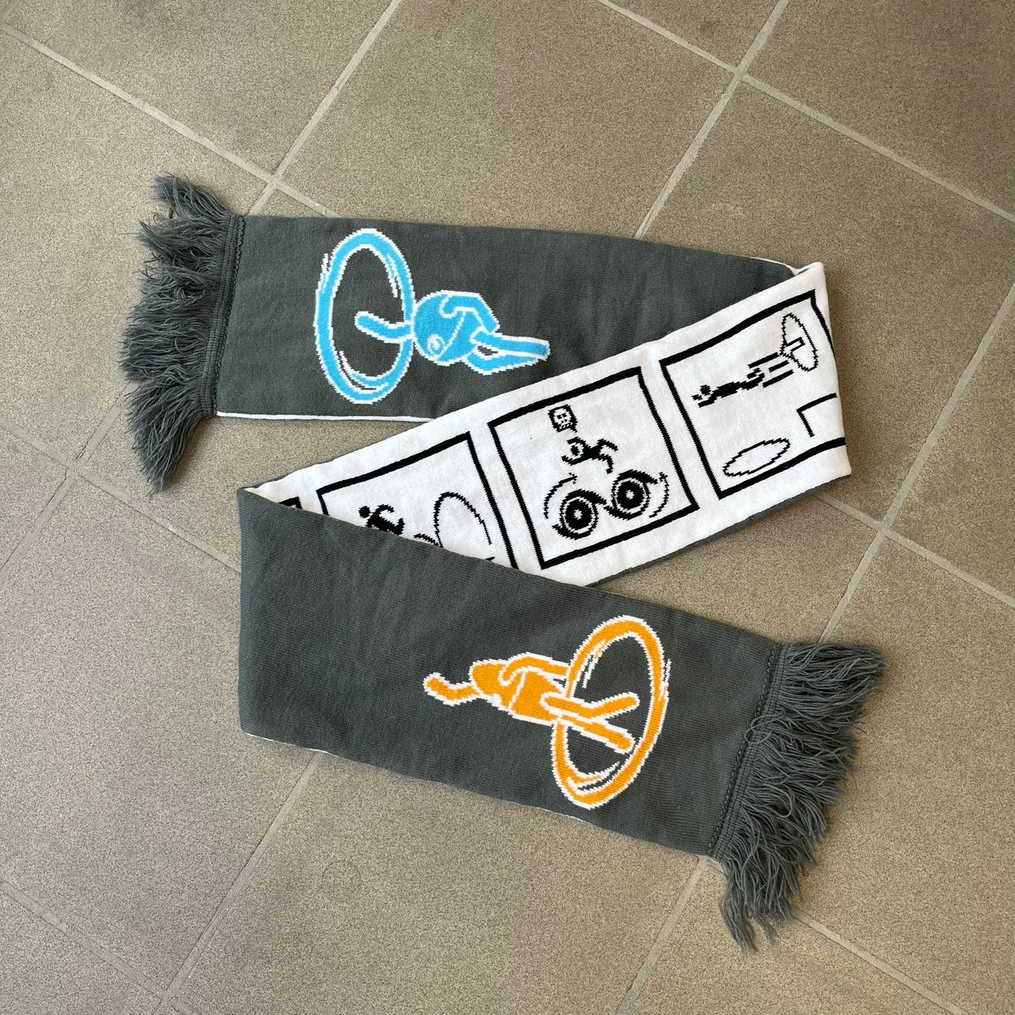 Portal 2 Chamber Squares 2015 Knit Scarf features a reversible design. One side is grey with an Aperture Laboratories logo in the center, along with two portals containing Atlas and P-body, one blue and one orange. The other side is white with a repeating pattern of Portal 2 chamber squares. Made by The Coop in 2015.
