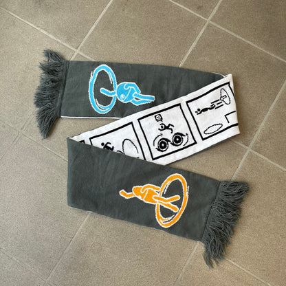 Portal 2 Chamber Squares 2015 Knit Scarf features a reversible design. One side is grey with an Aperture Laboratories logo in the center, along with two portals containing Atlas and P-body, one blue and one orange. The other side is white with a repeating pattern of Portal 2 chamber squares. Made by The Coop in 2015.