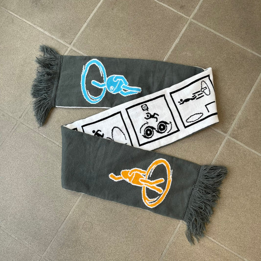 Portal 2 Chamber Squares 2015 Knit Scarf features a reversible design. One side is grey with an Aperture Laboratories logo in the center, along with two portals containing Atlas and P-body, one blue and one orange. The other side is white with a repeating pattern of Portal 2 chamber squares. Made by The Coop in 2015.
