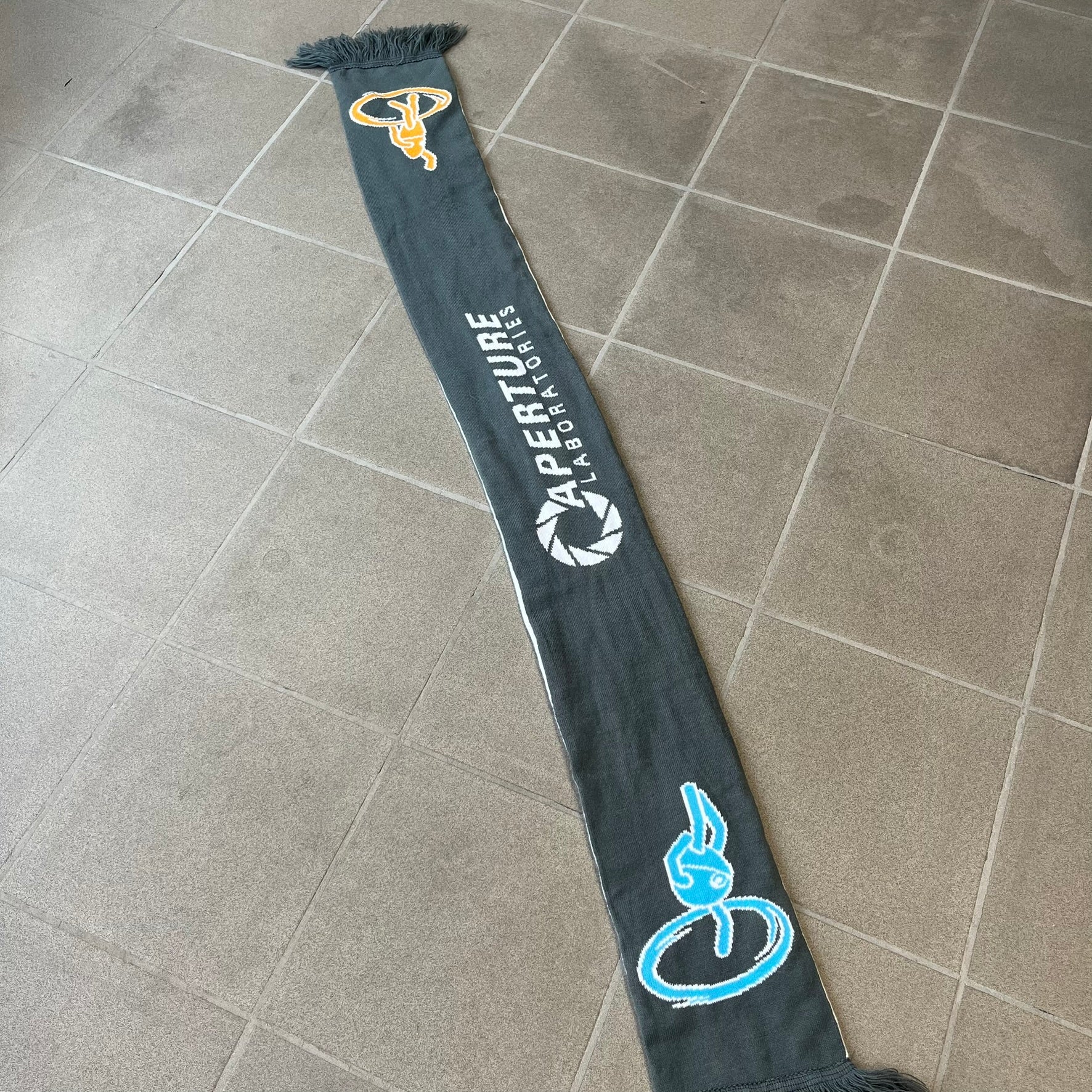 Portal 2 Chamber Squares 2015 Knit Scarf features a reversible design. One side is grey with an Aperture Laboratories logo in the center, along with two portals containing Atlas and P-body, one blue and one orange. The other side is white with a repeating pattern of Portal 2 chamber squares. Made by The Coop in 2015.