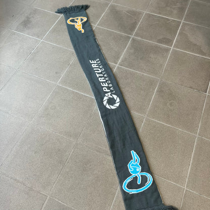 Portal 2 Chamber Squares 2015 Knit Scarf features a reversible design. One side is grey with an Aperture Laboratories logo in the center, along with two portals containing Atlas and P-body, one blue and one orange. The other side is white with a repeating pattern of Portal 2 chamber squares. Made by The Coop in 2015.