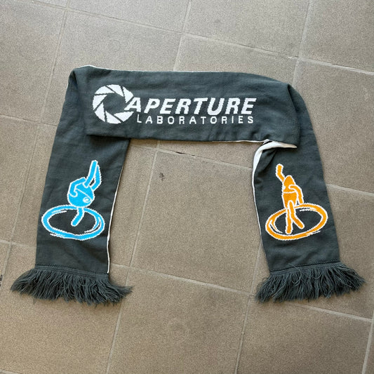 Portal 2 Chamber Squares 2015 Knit Scarf features a reversible design. One side is grey with an Aperture Laboratories logo in the center, along with two portals containing Atlas and P-body, one blue and one orange. The other side is white with a repeating pattern of Portal 2 chamber squares. Made by The Coop in 2015.