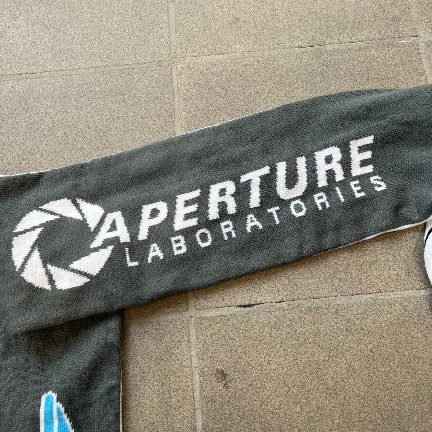 Portal 2 Chamber Squares 2015 Knit Scarf features a reversible design. One side is grey with an Aperture Laboratories logo in the center, along with two portals containing Atlas and P-body, one blue and one orange. The other side is white with a repeating pattern of Portal 2 chamber squares. Made by The Coop in 2015.
