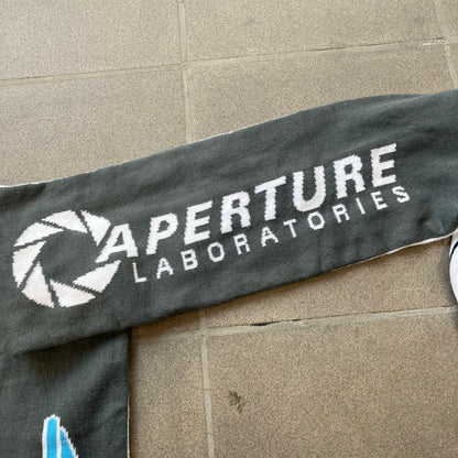 Portal 2 Chamber Squares 2015 Knit Scarf features a reversible design. One side is grey with an Aperture Laboratories logo in the center, along with two portals containing Atlas and P-body, one blue and one orange. The other side is white with a repeating pattern of Portal 2 chamber squares. Made by The Coop in 2015.