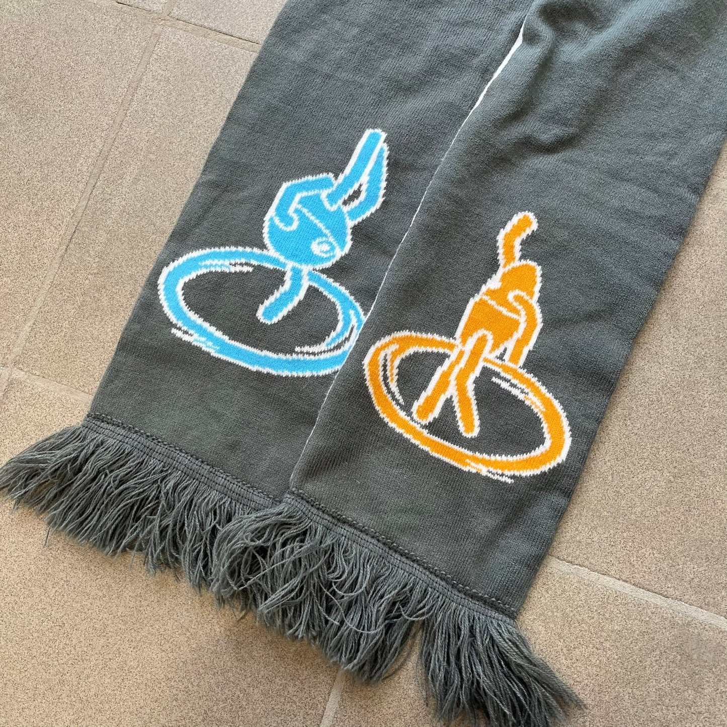 Portal 2 Chamber Squares 2015 Knit Scarf features a reversible design. One side is grey with an Aperture Laboratories logo in the center, along with two portals containing Atlas and P-body, one blue and one orange. The other side is white with a repeating pattern of Portal 2 chamber squares. Made by The Coop in 2015.