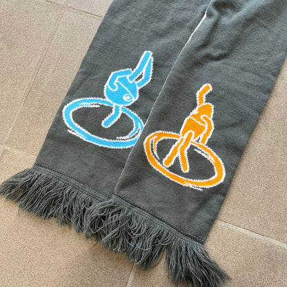 Portal 2 Chamber Squares 2015 Knit Scarf features a reversible design. One side is grey with an Aperture Laboratories logo in the center, along with two portals containing Atlas and P-body, one blue and one orange. The other side is white with a repeating pattern of Portal 2 chamber squares. Made by The Coop in 2015.