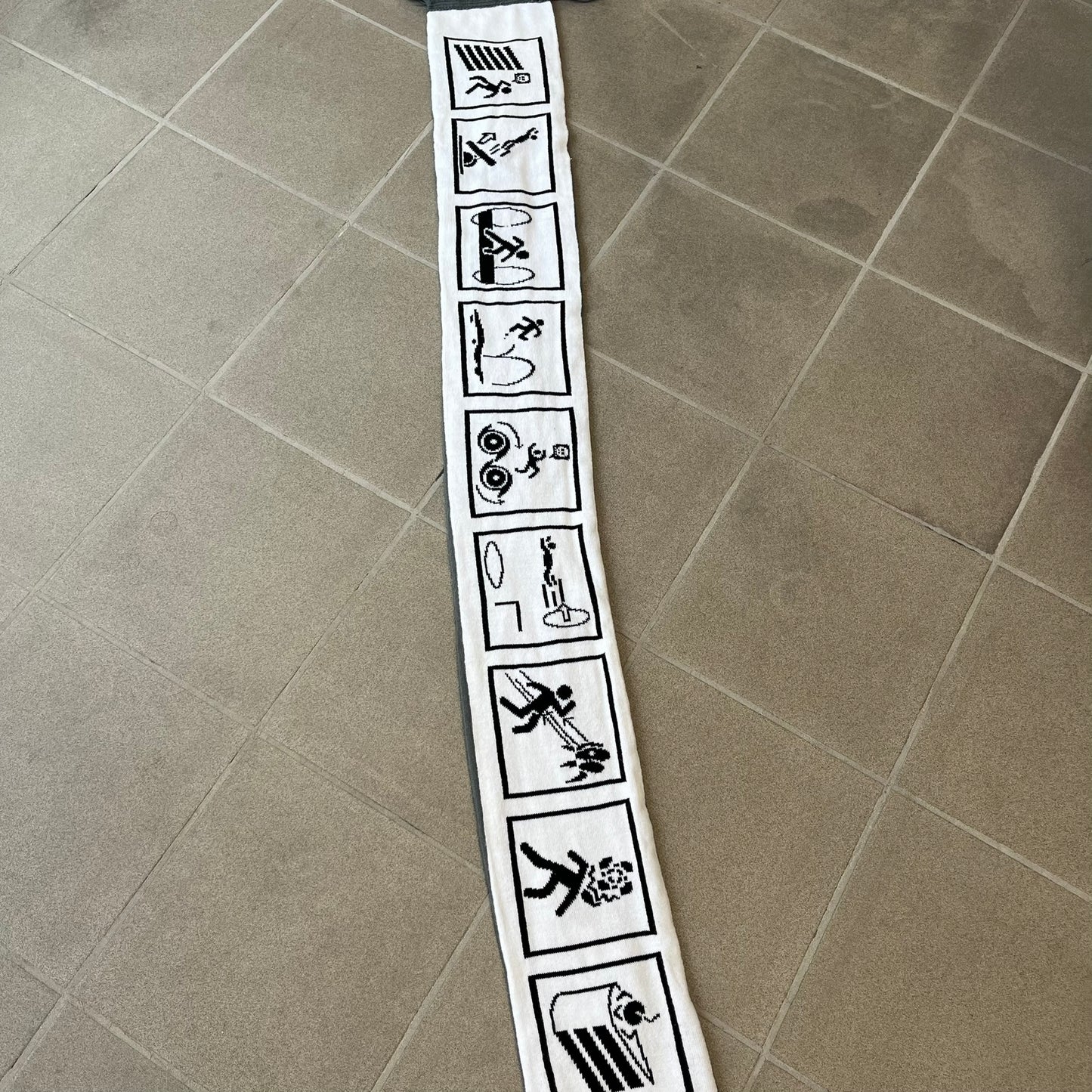 Portal 2 Chamber Squares 2015 Knit Scarf features a reversible design. One side is grey with an Aperture Laboratories logo in the center, along with two portals containing Atlas and P-body, one blue and one orange. The other side is white with a repeating pattern of Portal 2 chamber squares. Made by The Coop in 2015.