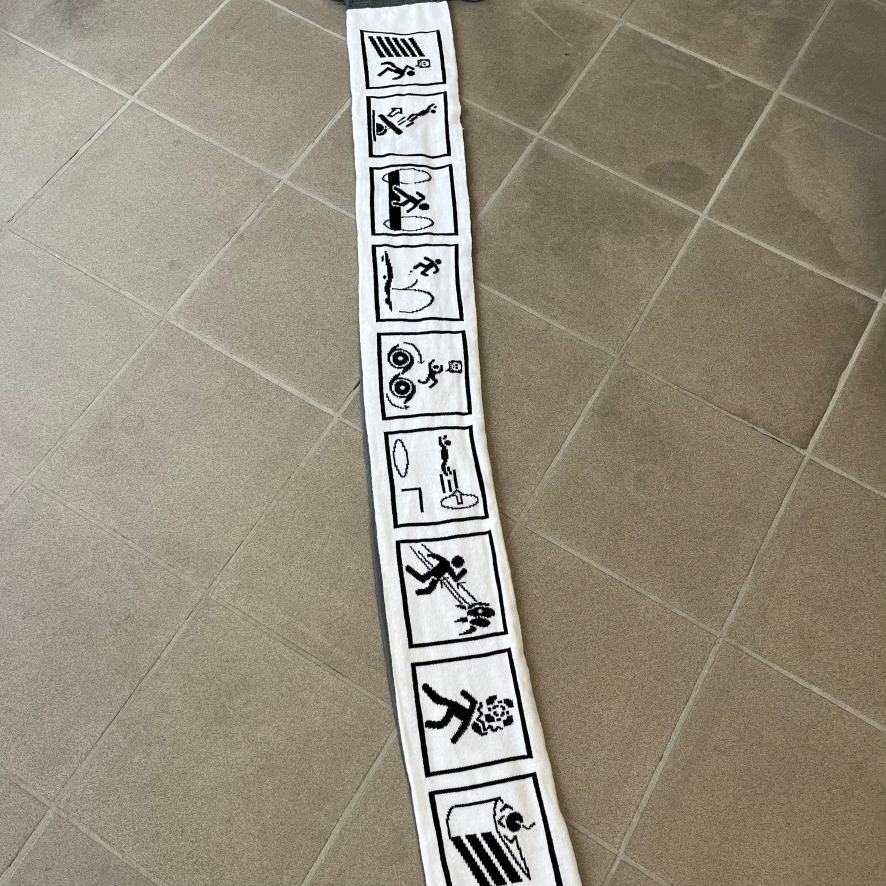 Portal 2 Chamber Squares 2015 Knit Scarf features a reversible design. One side is grey with an Aperture Laboratories logo in the center, along with two portals containing Atlas and P-body, one blue and one orange. The other side is white with a repeating pattern of Portal 2 chamber squares. Made by The Coop in 2015.
