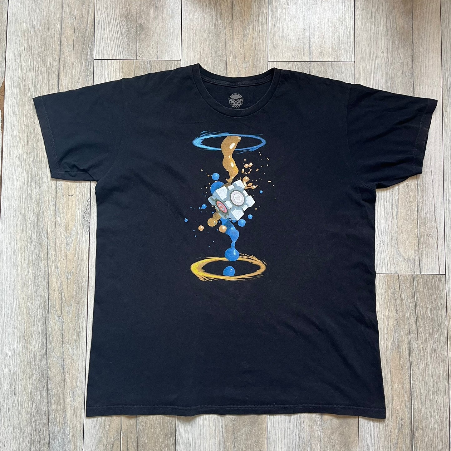 Portal 2 Companion Cube 2016 J!NX Tee is a black T-shirt produced by J!NX in collaboration with Valve. The front features a Companion Cube graphic centered between a blue portal at the top and an orange portal at the bottom, with contrasting colored flow effects connecting and splashing onto the cube. Small Valve licensing text appears beneath the lower portal.