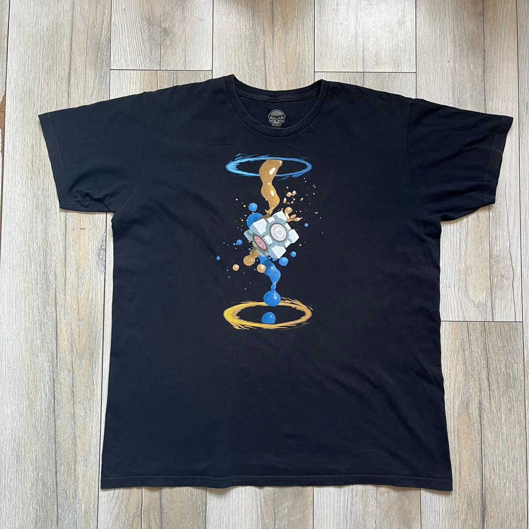 Portal 2 Companion Cube 2016 J!NX Tee is a black T-shirt produced by J!NX in collaboration with Valve. The front features a Companion Cube graphic centered between a blue portal at the top and an orange portal at the bottom, with contrasting colored flow effects connecting and splashing onto the cube. Small Valve licensing text appears beneath the lower portal.