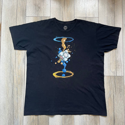 Portal 2 Companion Cube 2016 J!NX Tee is a black T-shirt produced by J!NX in collaboration with Valve. The front features a Companion Cube graphic centered between a blue portal at the top and an orange portal at the bottom, with contrasting colored flow effects connecting and splashing onto the cube. Small Valve licensing text appears beneath the lower portal.