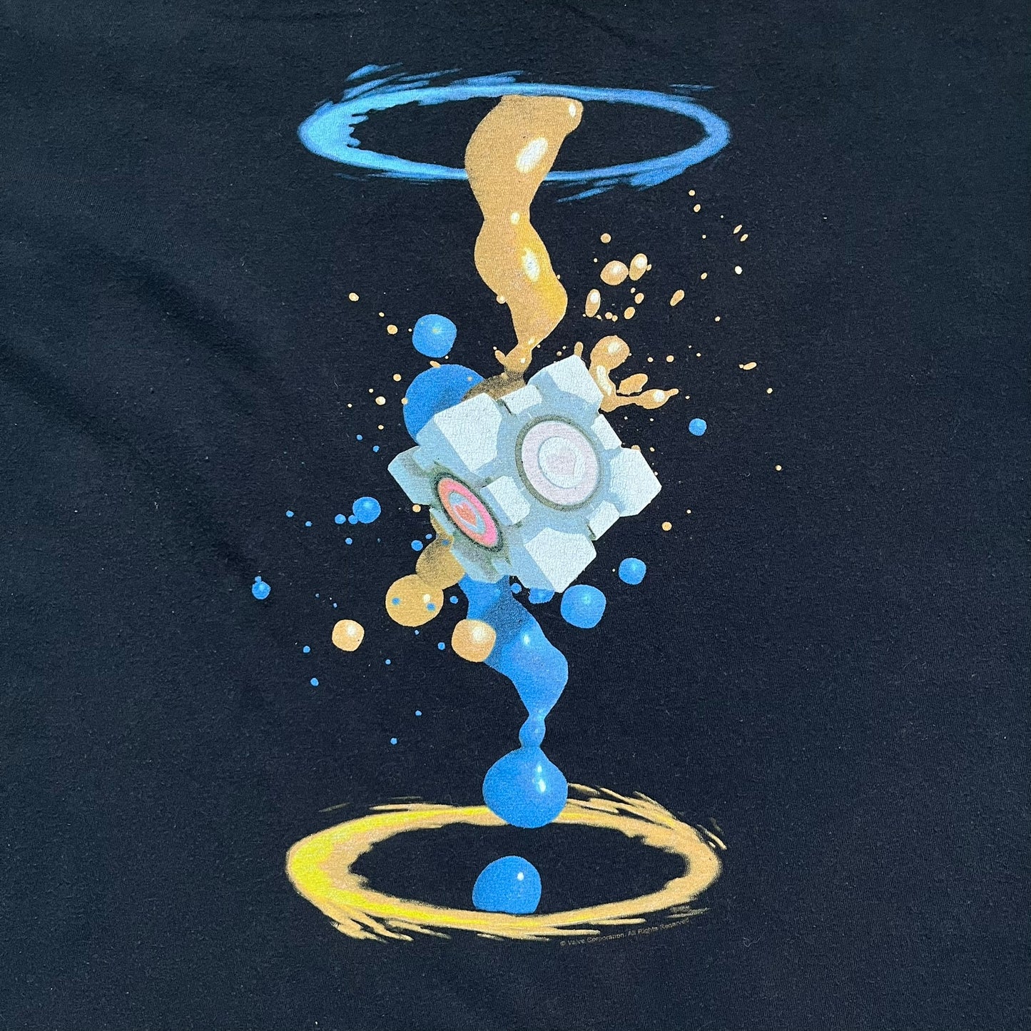 Portal 2 Companion Cube 2016 J!NX Tee is a black T-shirt produced by J!NX in collaboration with Valve. The front features a Companion Cube graphic centered between a blue portal at the top and an orange portal at the bottom, with contrasting colored flow effects connecting and splashing onto the cube. Small Valve licensing text appears beneath the lower portal.