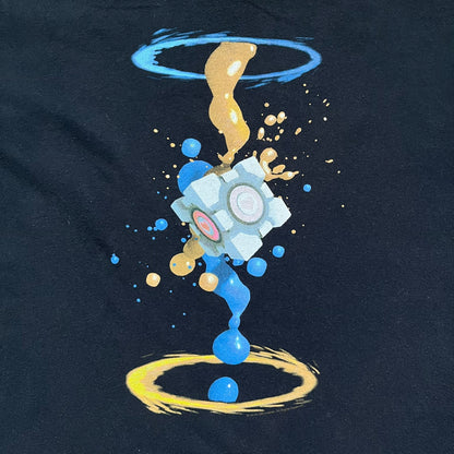 Portal 2 Companion Cube 2016 J!NX Tee is a black T-shirt produced by J!NX in collaboration with Valve. The front features a Companion Cube graphic centered between a blue portal at the top and an orange portal at the bottom, with contrasting colored flow effects connecting and splashing onto the cube. Small Valve licensing text appears beneath the lower portal.