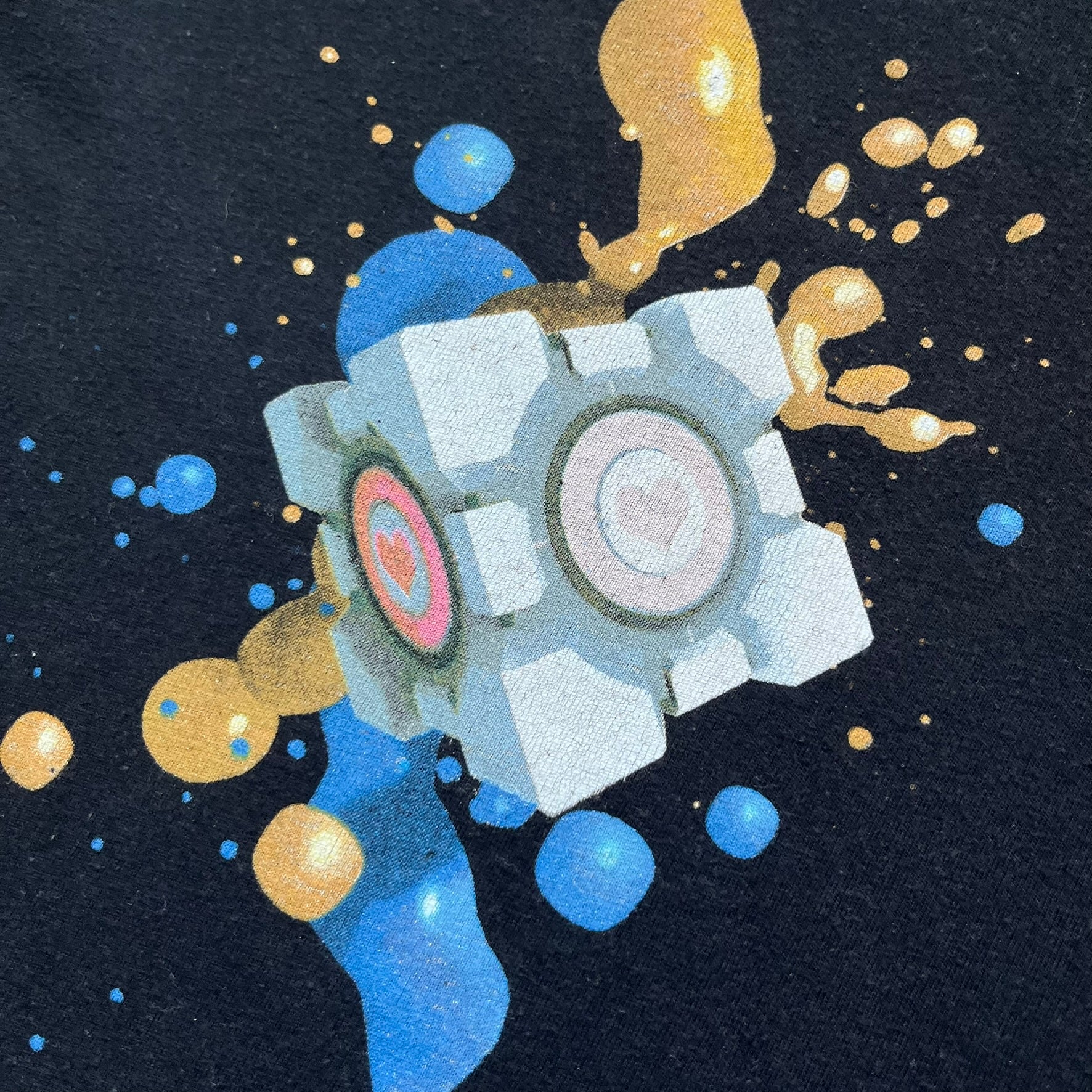 Portal 2 Companion Cube 2016 J!NX Tee is a black T-shirt produced by J!NX in collaboration with Valve. The front features a Companion Cube graphic centered between a blue portal at the top and an orange portal at the bottom, with contrasting colored flow effects connecting and splashing onto the cube. Small Valve licensing text appears beneath the lower portal.
