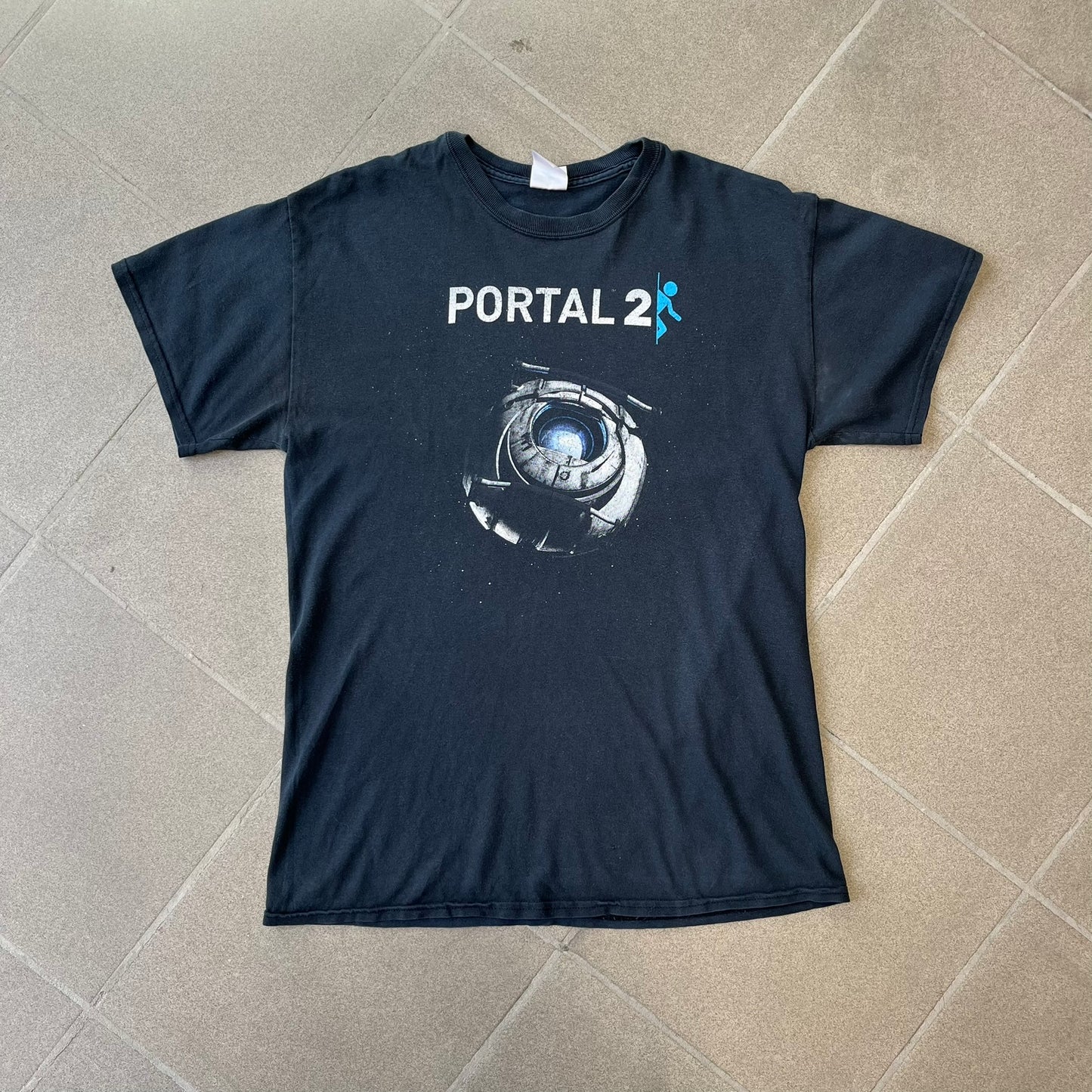 Portal 2: Wheatley in Space 2011 J!nx Tee features a black T-shirt made on a Gildan blank by J!NX in collaboration with Valve. The front has a graphic of Wheatley floating in space with a white and blue Portal 2 logo above him. Made from 100% cotton.
